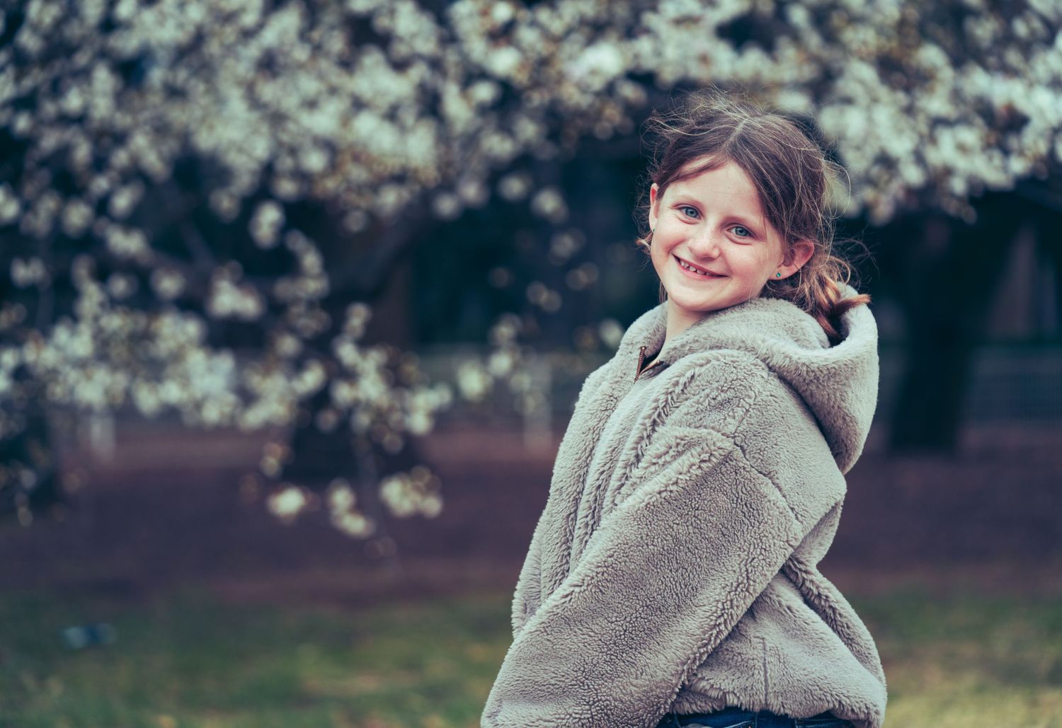 Child in cozy sweater smiles near flowering trees in spring.