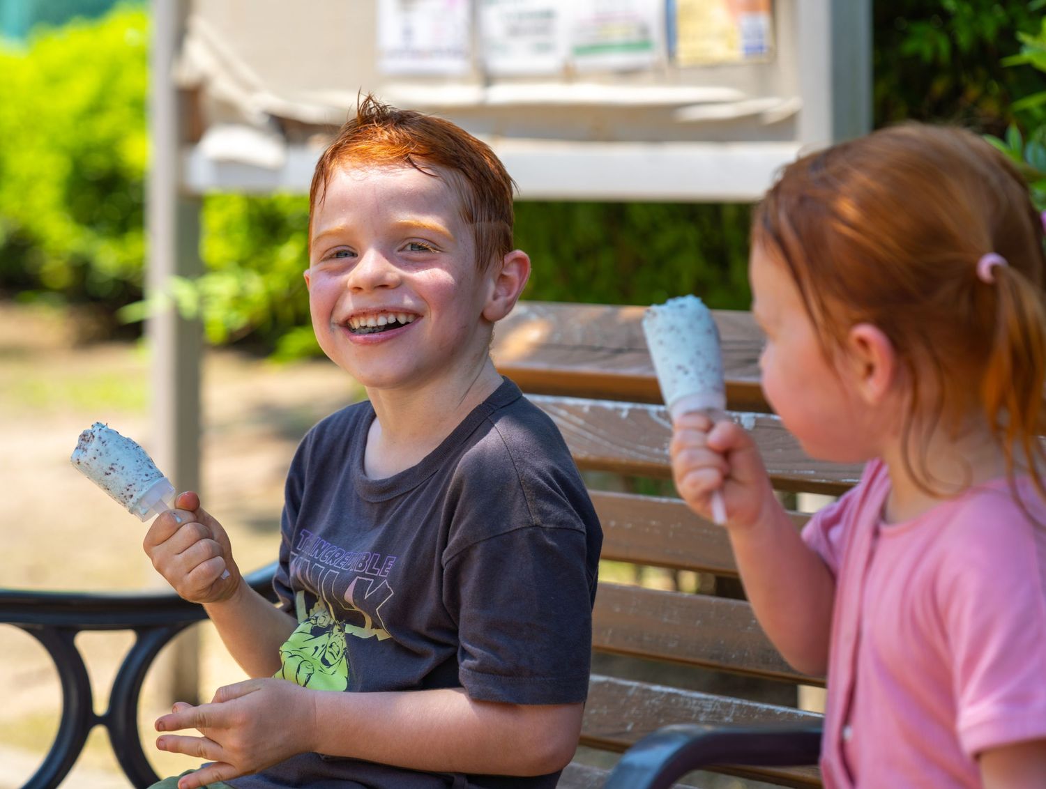 Children sit together at a picnic table outdoors enjoying frozen treats on a sunny day.