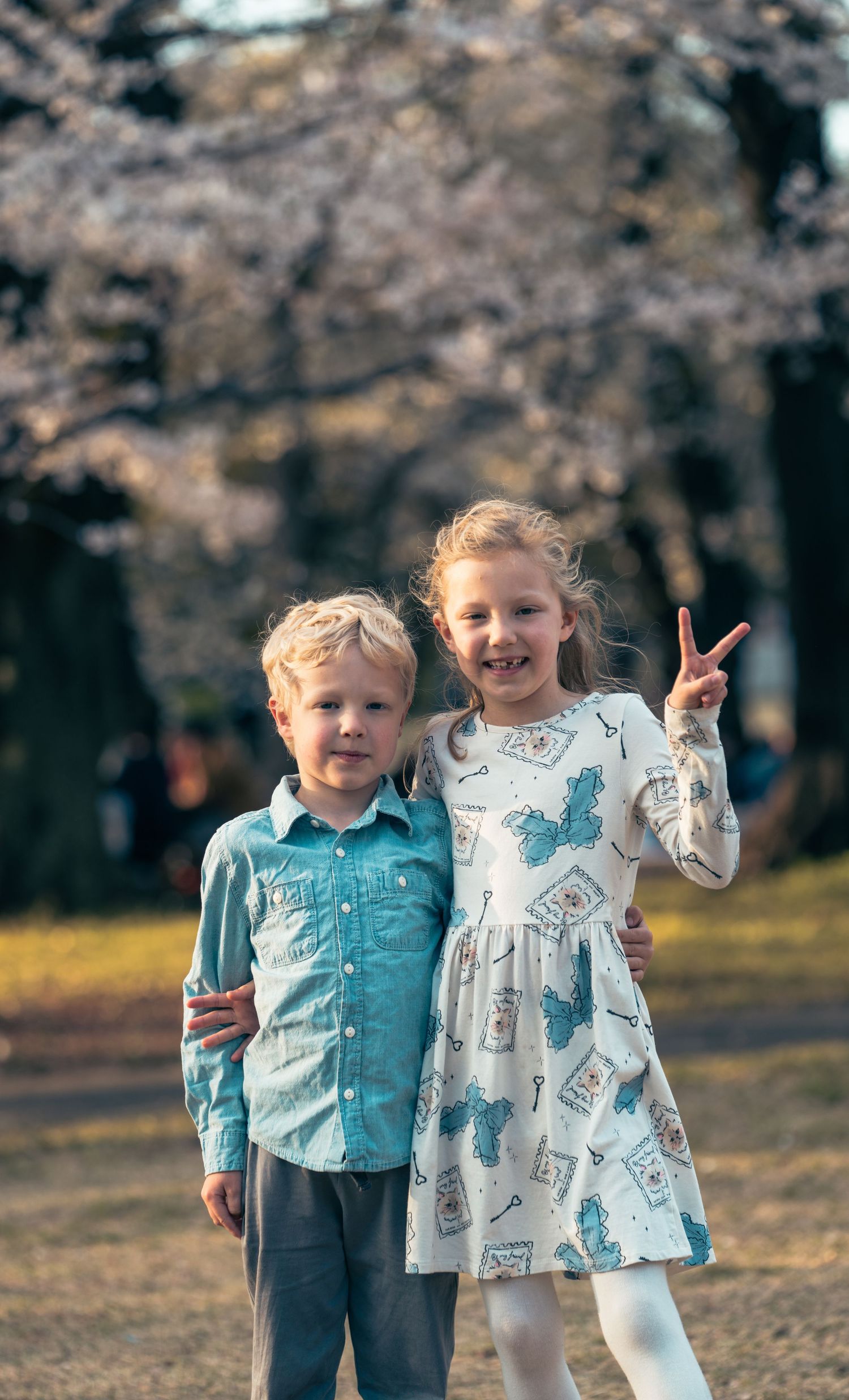 Two children standing together under cherry blossoms with one making a peace sign.
