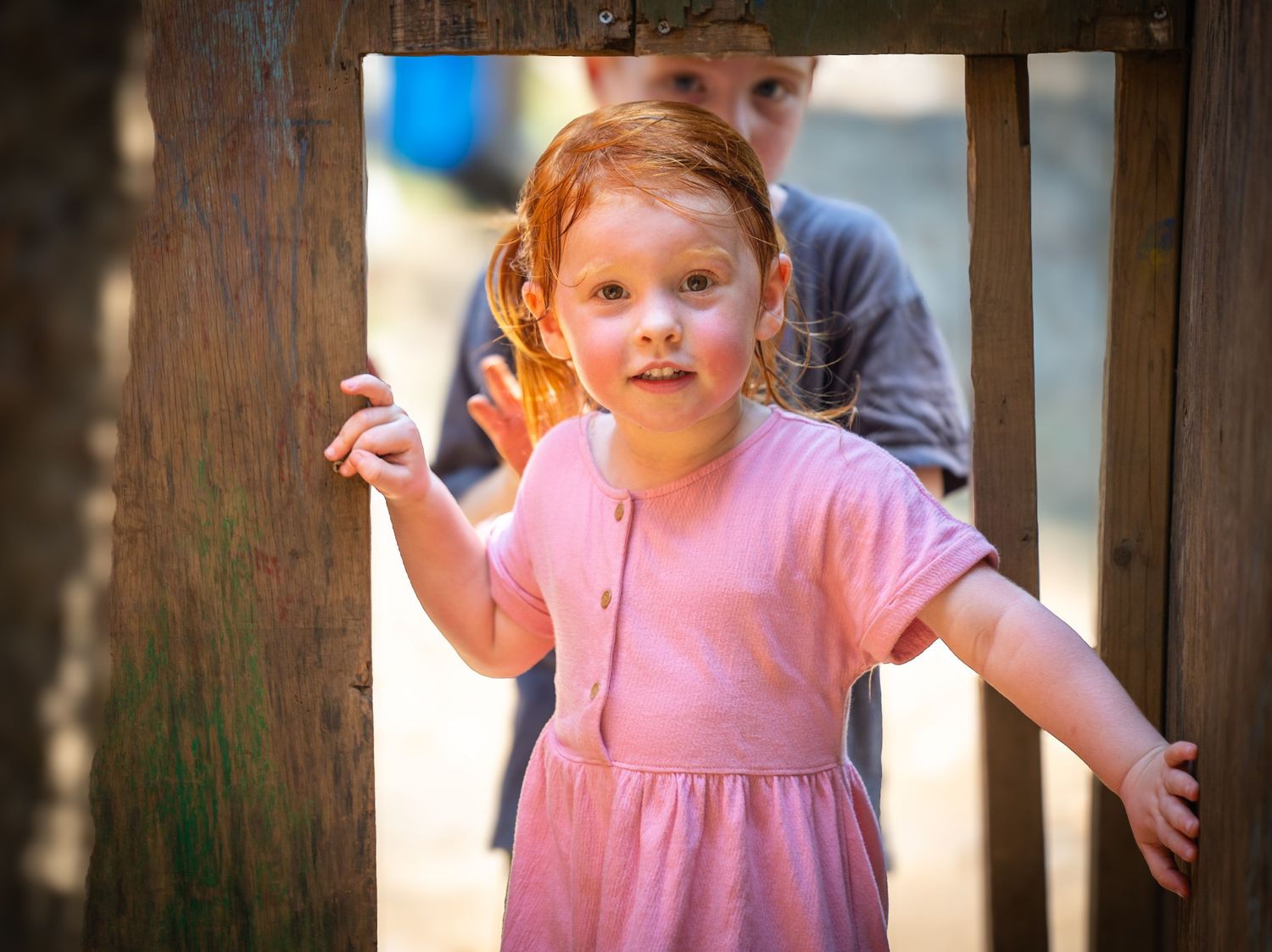 A child in a pink dress stands near a wooden fence post with arms outstretched while playing outdoors.