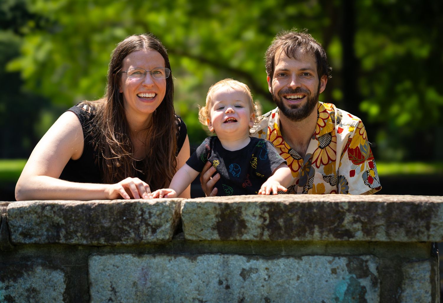 Three people lean against a stone wall outdoors on a sunny day, smiling together in a natural setting.