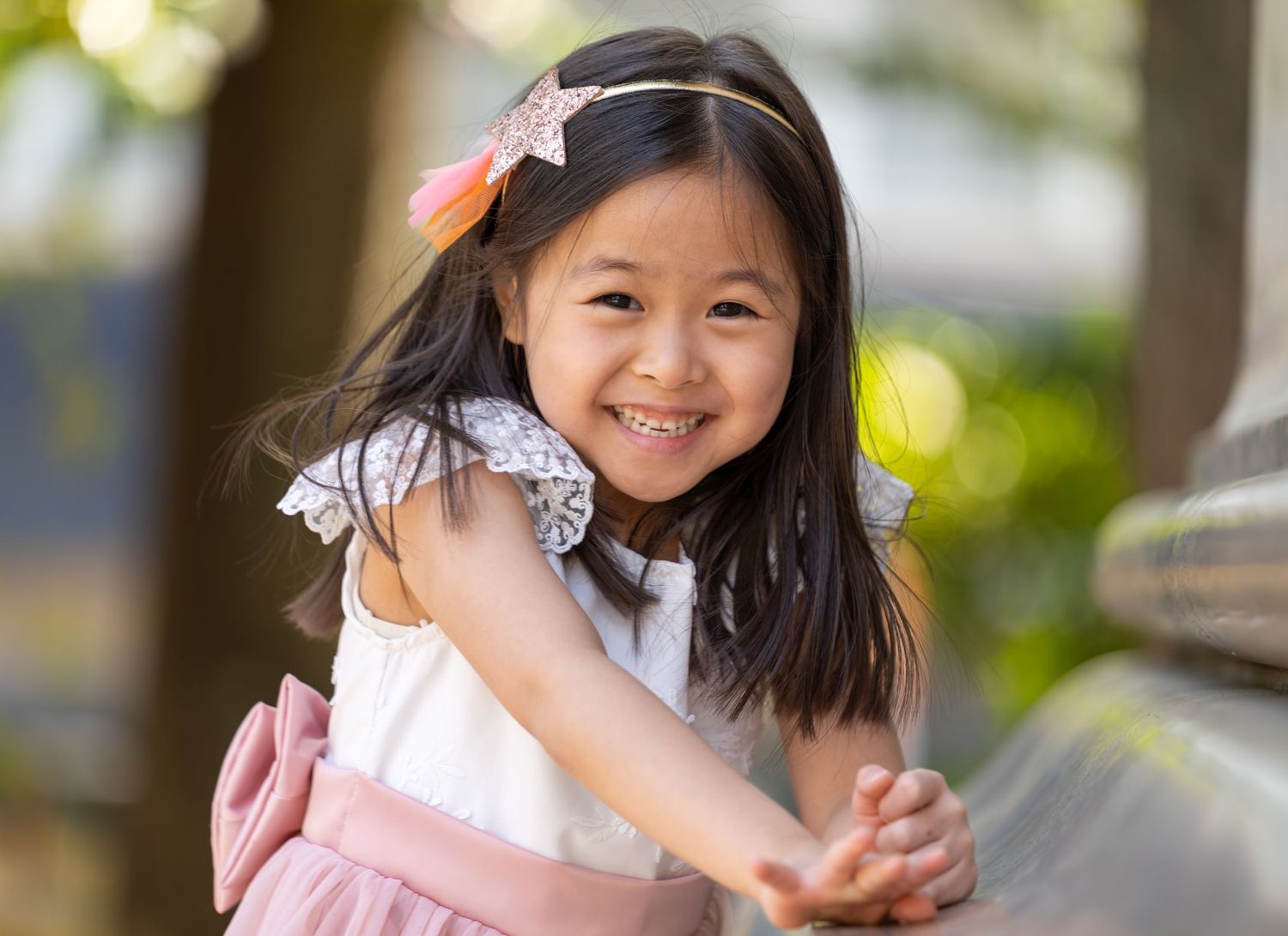 A smiling young Asian girl wearing a white dress and pink bow headband, leaning forward outdoors.