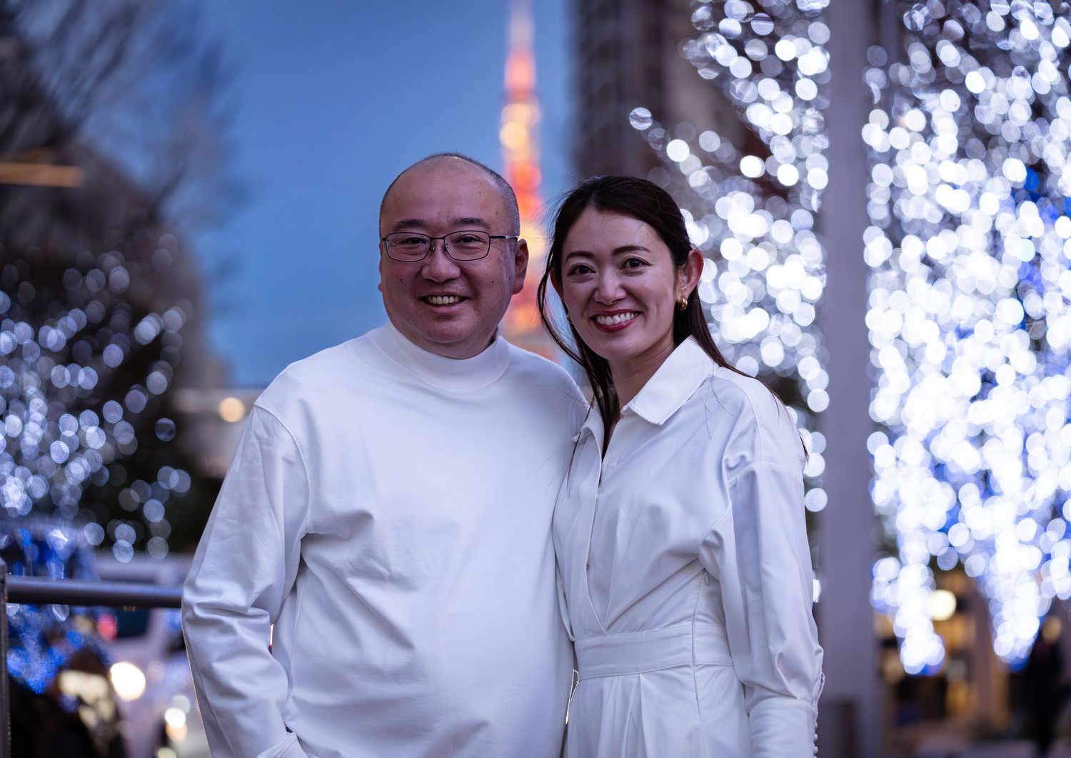 A smiling couple in matching white outfits pose in front of sparkling bokeh lights with Tokyo Tower at dusk.