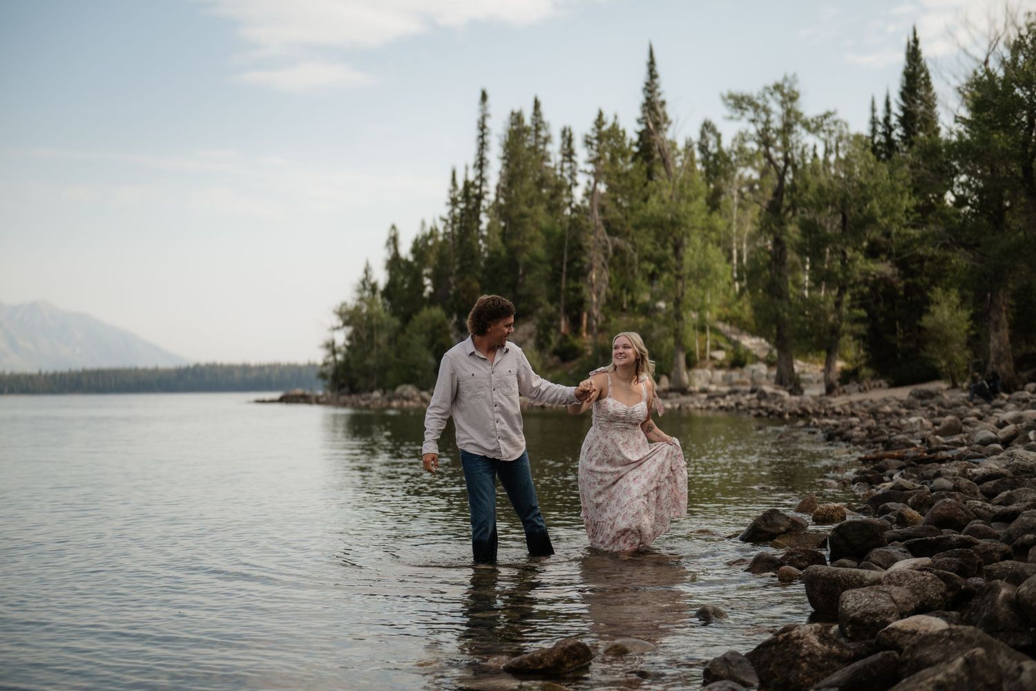 A couple wades in the shallow waters of a picturesque lake surrounded by a forested landscape.