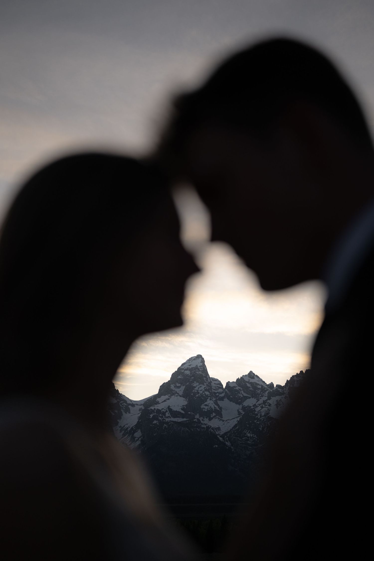 Silhouette of couple touching foreheads with snow-capped mountain peaks visible between them at dusk.