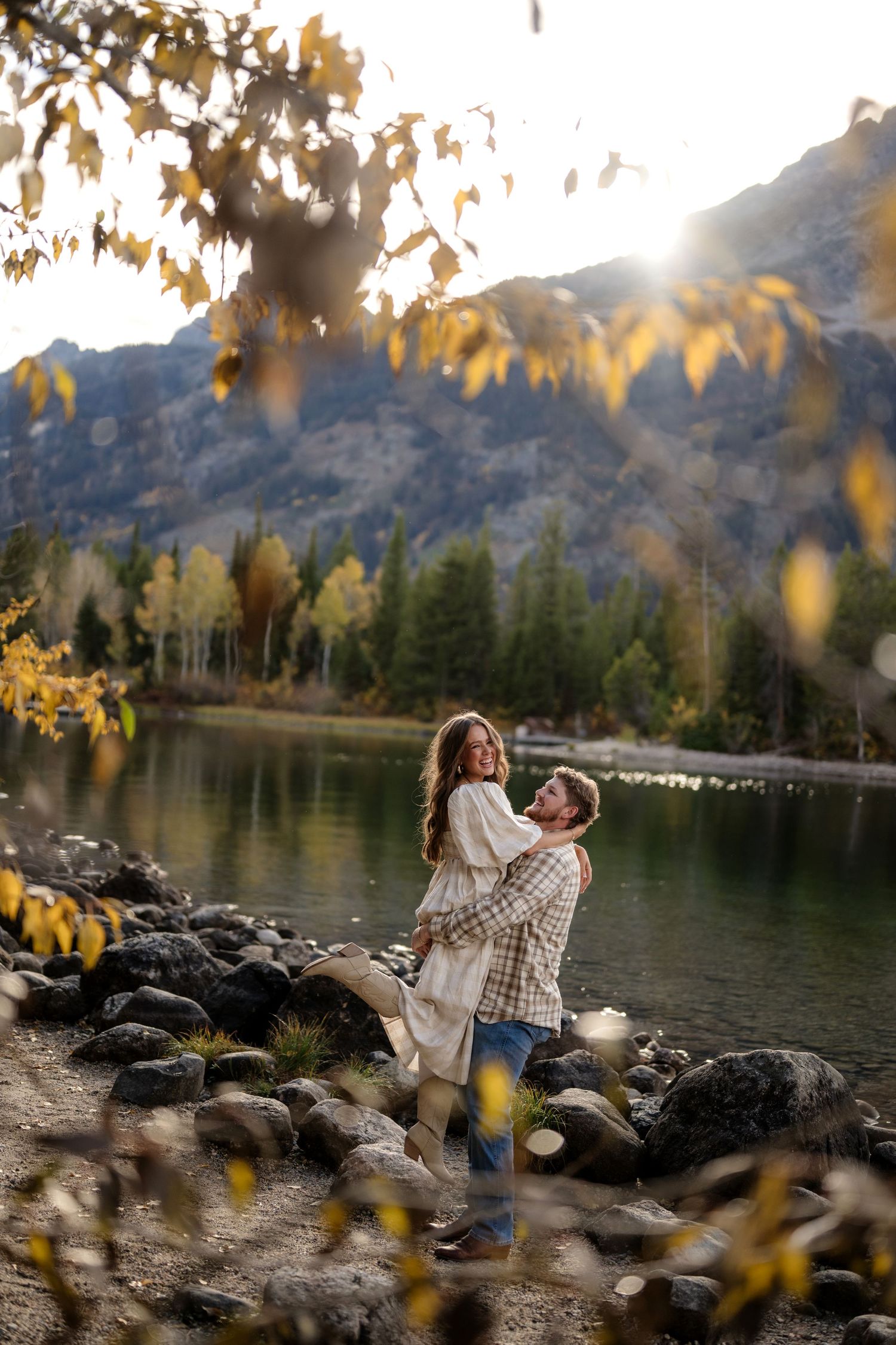 Couple embracing by a mountain lake in autumn, surrounded by golden leaves and evergreen trees at sunset.