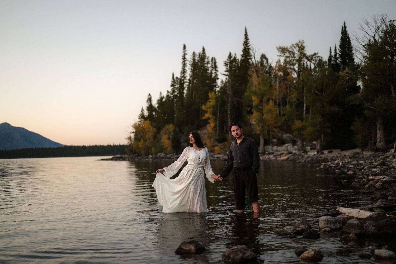 A couple stands waist-deep in a lake, embracing, with pine trees and mountains in the background at sunset.