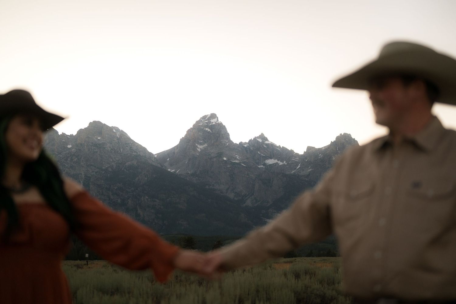 Two people hold hands in a grassy field with majestic mountains in the distance.