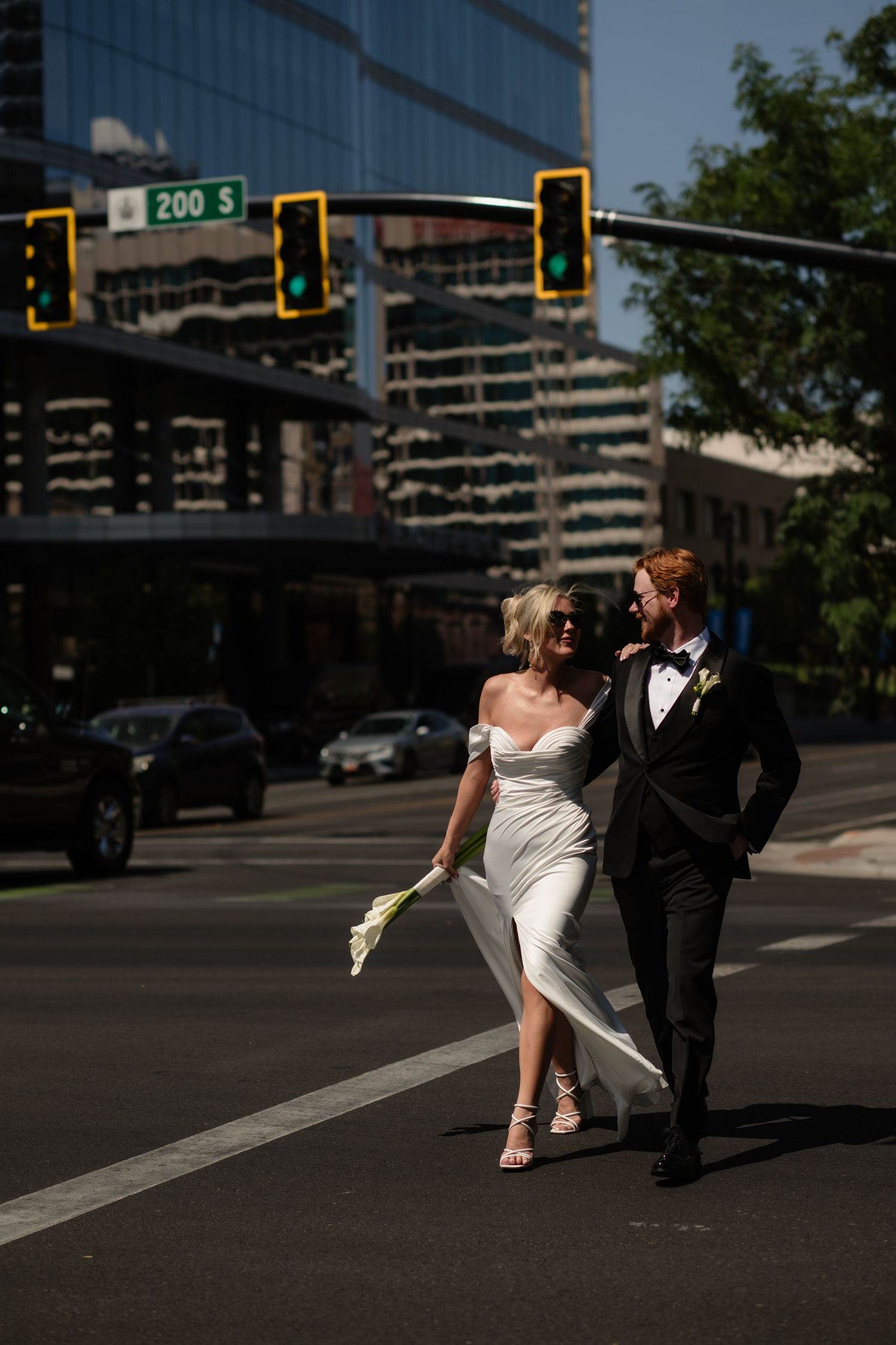 A bride and groom walk across a city street at night, with skyscrapers and traffic lights in the background.