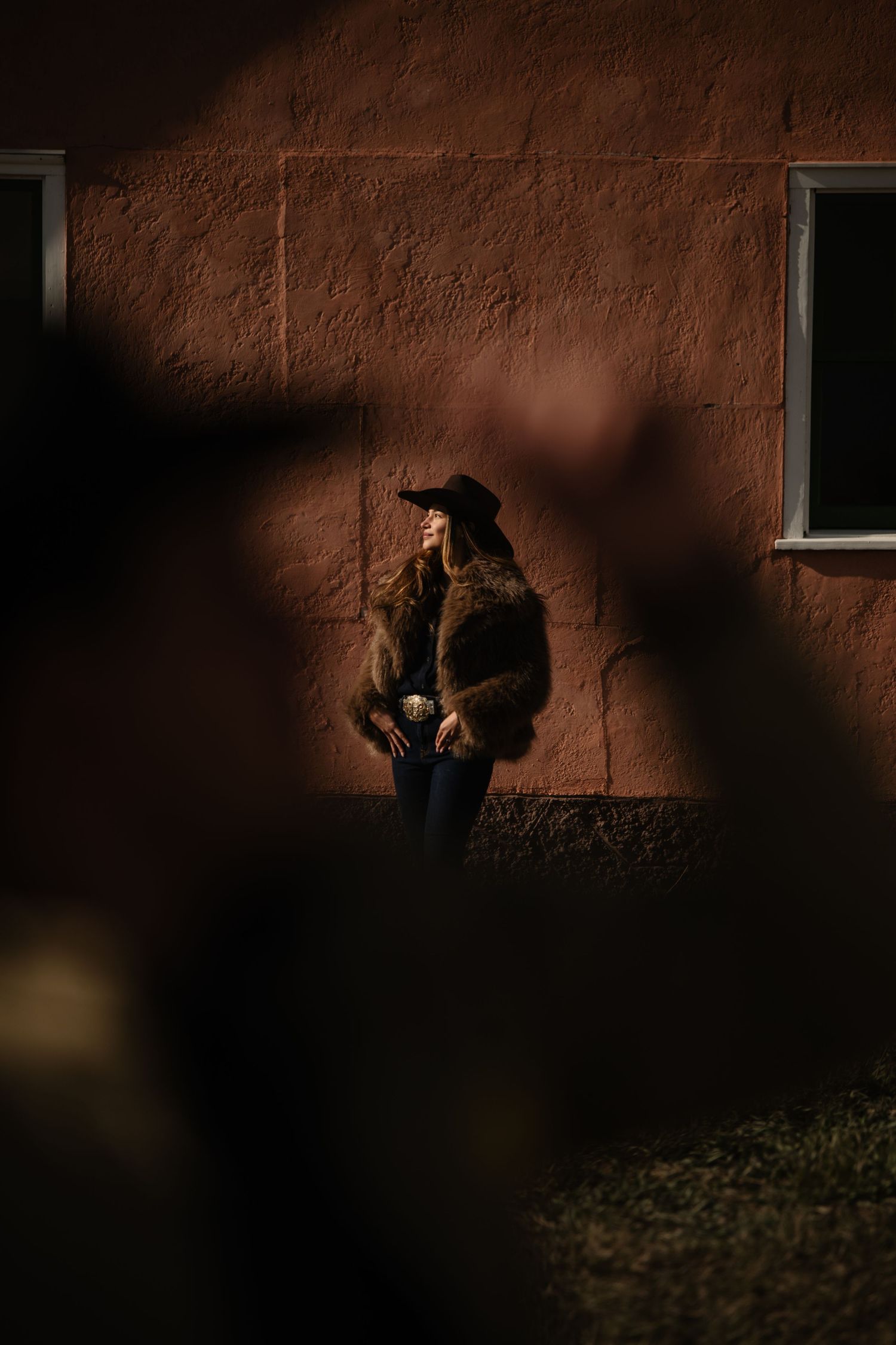Woman in fur coat and black hat leaning against a rustic red wall in dramatic sunlight.