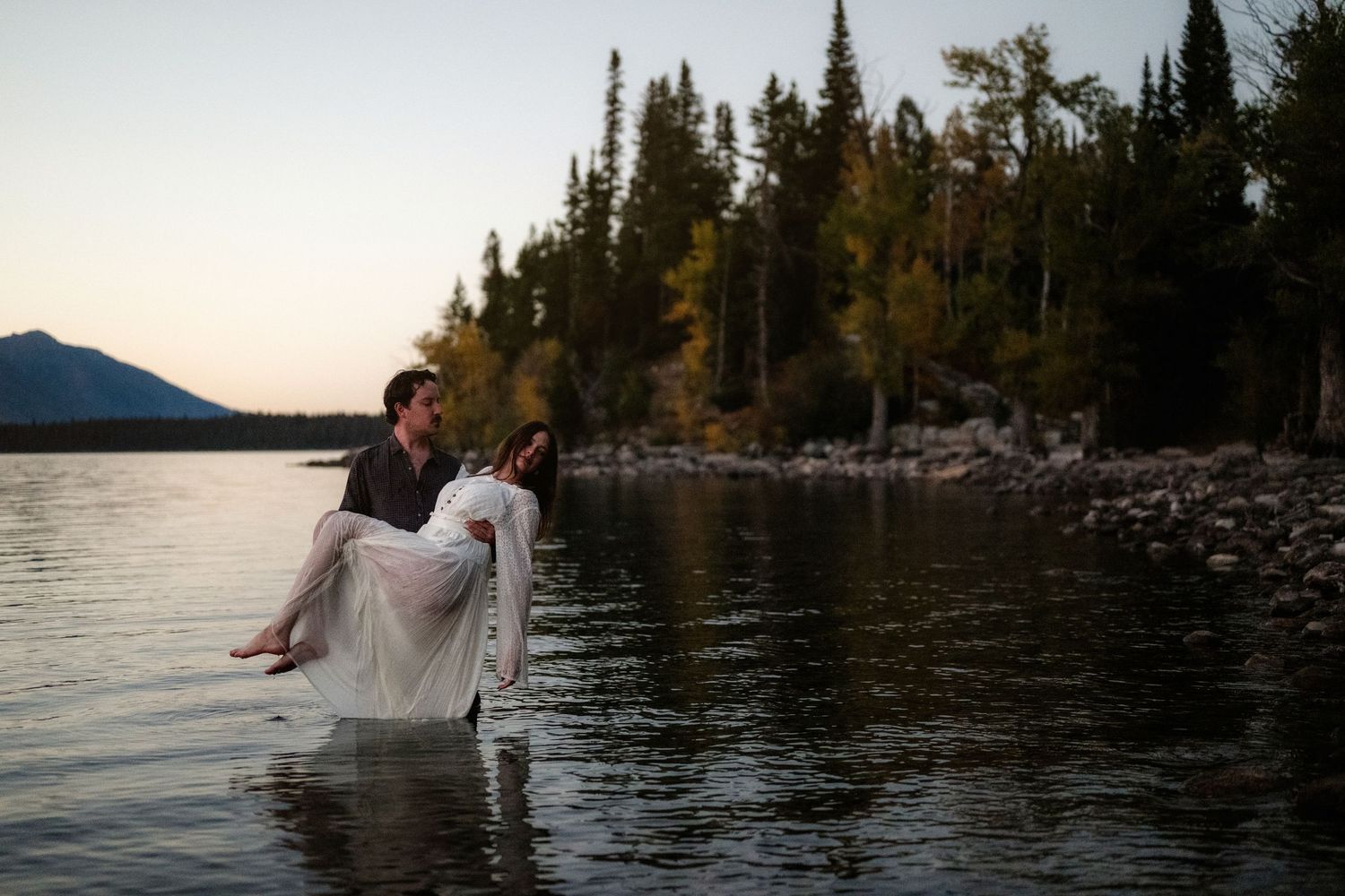 Groom carries bride in white dress through a serene mountain lake at dusk with evergreen trees in background.