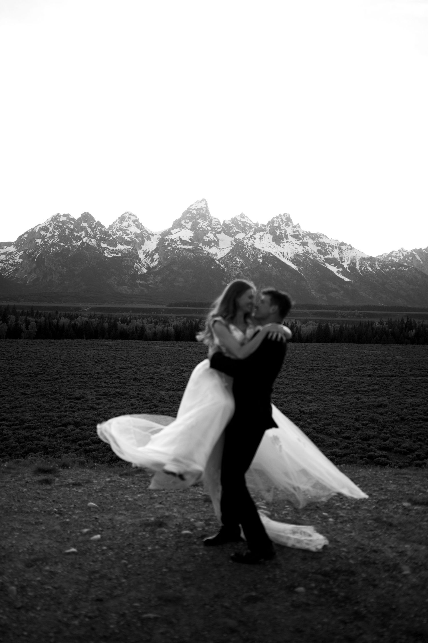 Black and white photo of groom lifting bride in wedding dress with Grand Teton mountains in background.