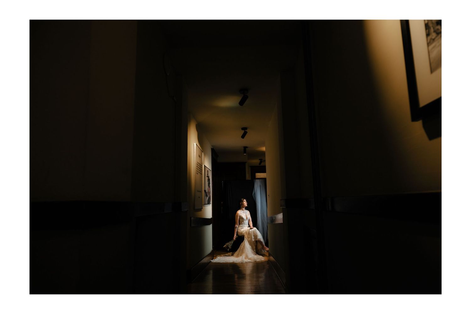 Bride in white gown sitting in a dimly lit hallway, creating a dramatic and elegant wedding portrait.