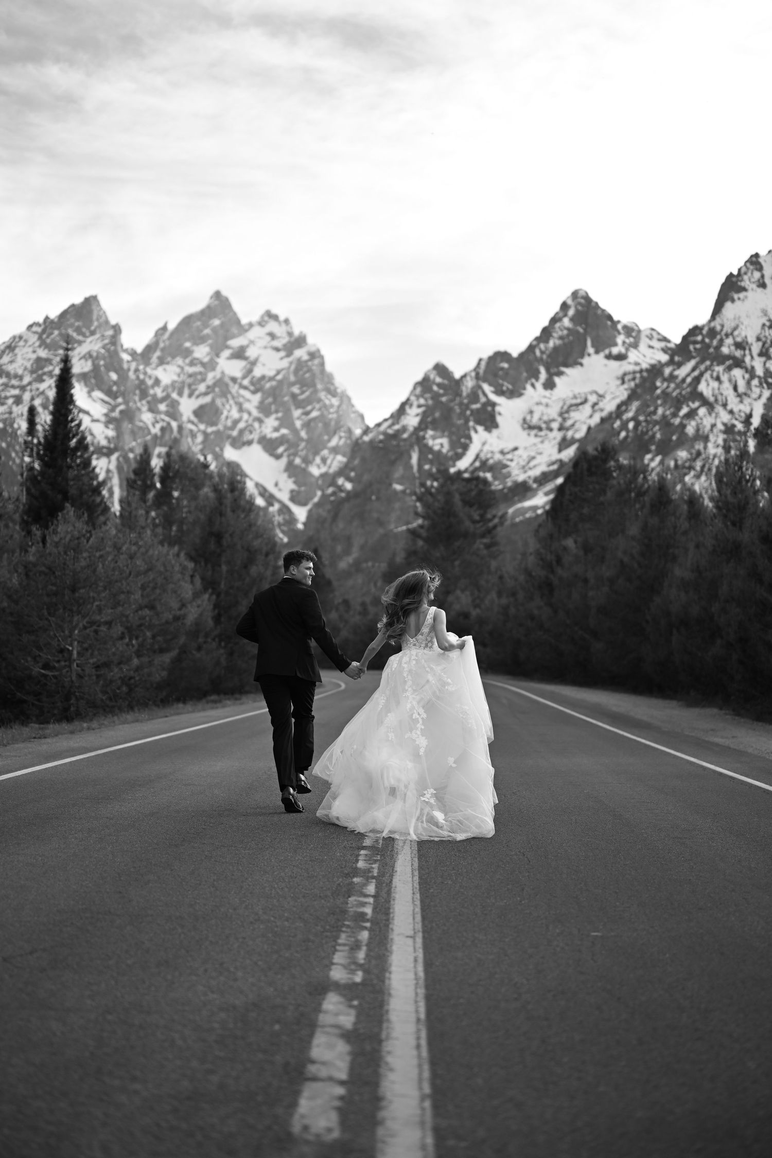 Black and white photo of bride and groom holding hands walking down a road toward snow-capped Grand Teton mountains.