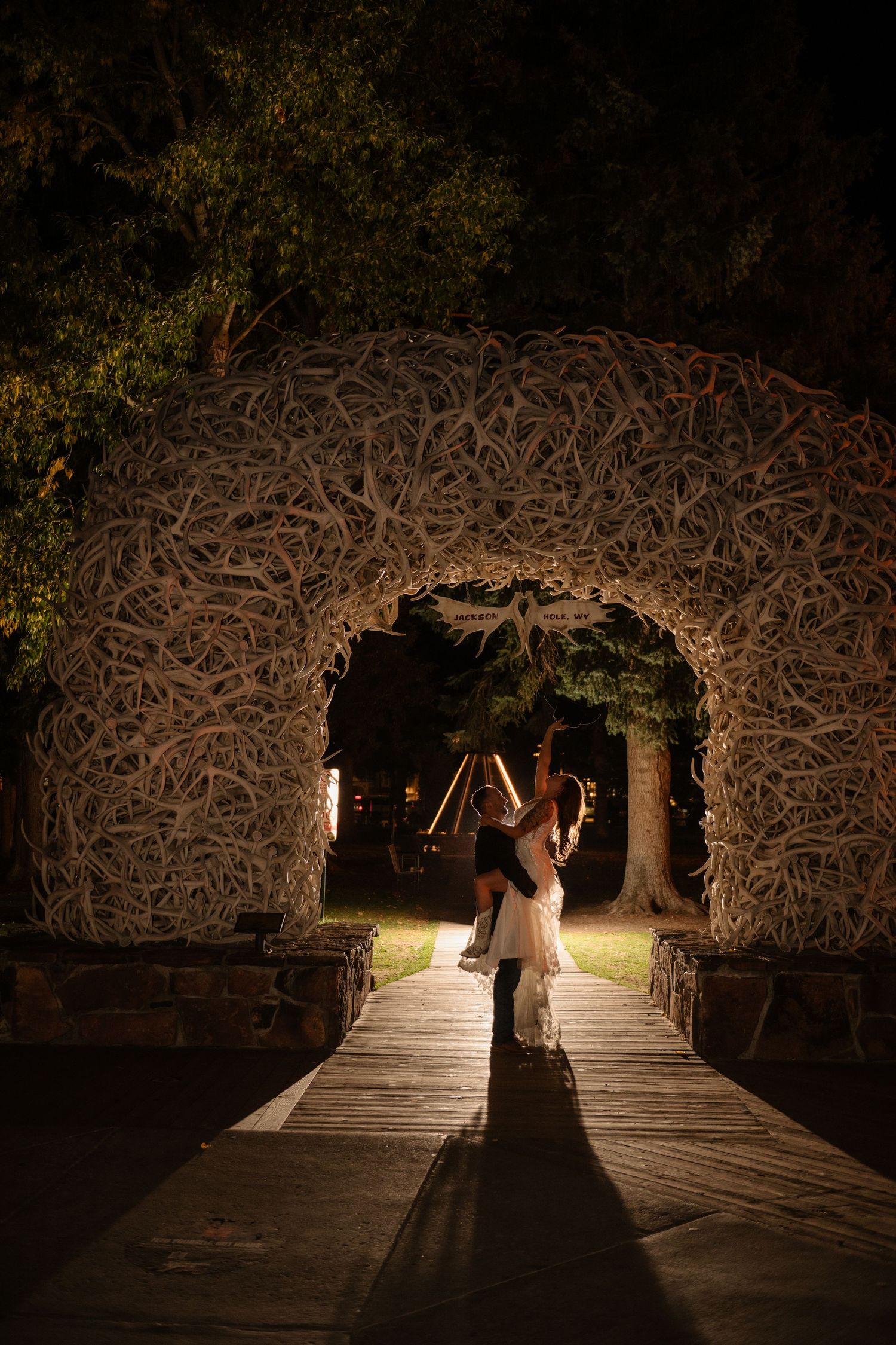 Couple dancing under illuminated antler arch on wooden walkway at night in romantic outdoor wedding setting.
