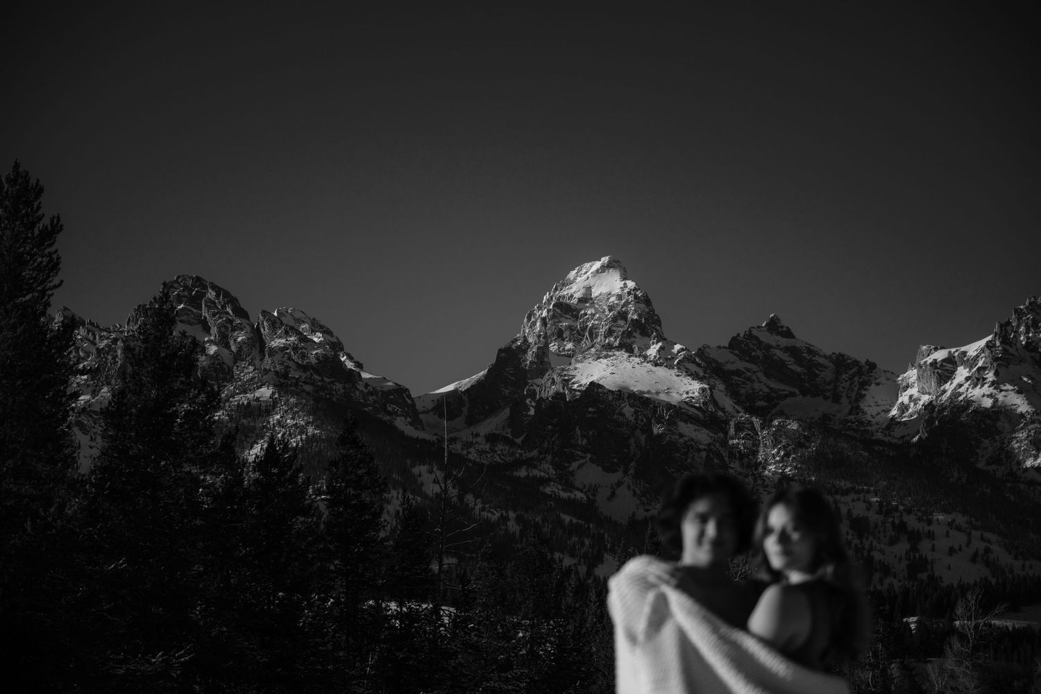 Black and white photo of a couple embracing before dramatic snow-capped Grand Teton mountains at dusk.