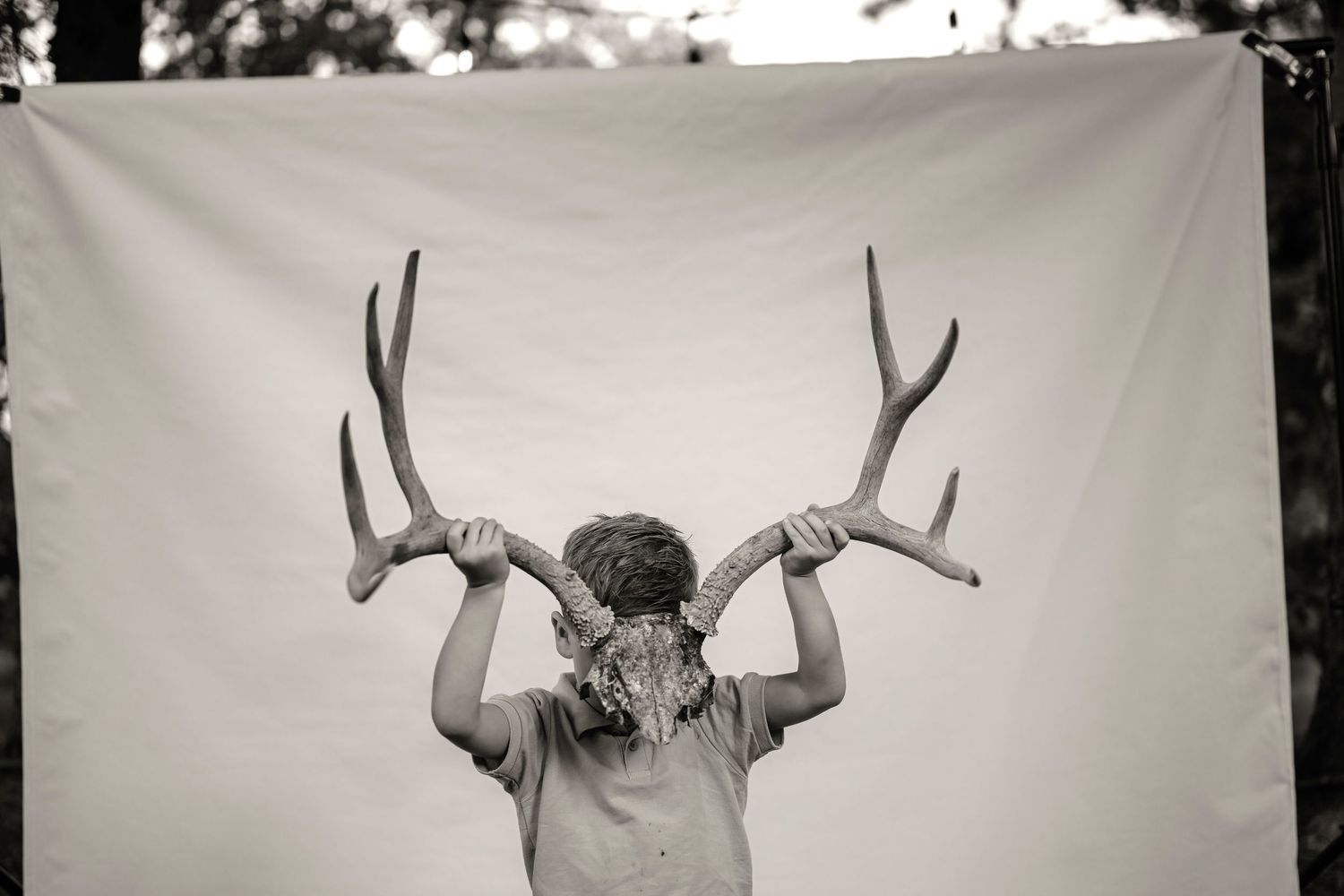 Child holding large deer antlers above their head against a white fabric backdrop in black and white photo.