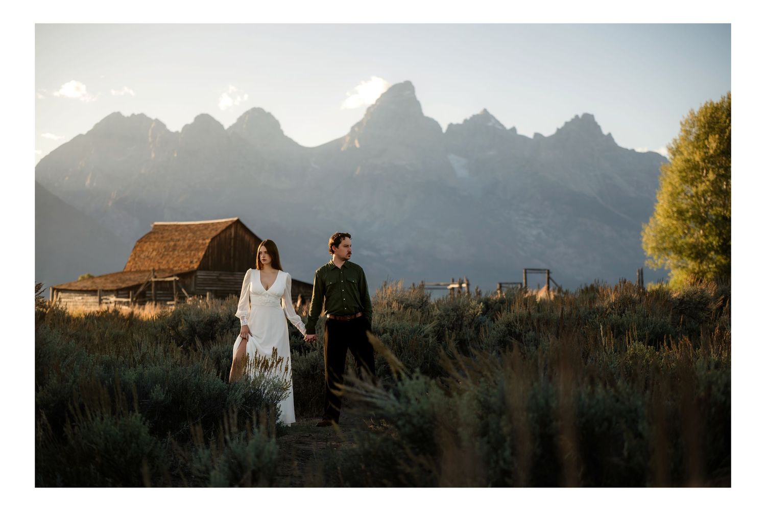 Couple walks hand-in-hand in a field with the Grand Teton mountains and a rustic barn in the background in Mormon Row Jackson Wyoming.