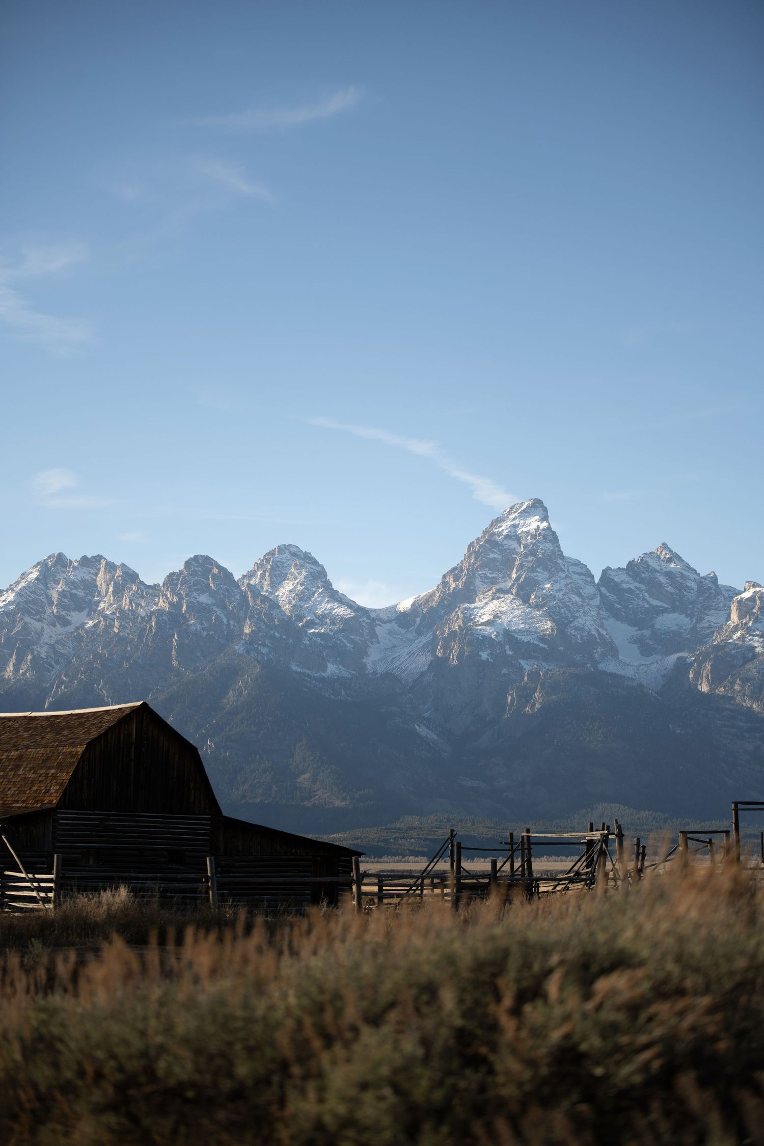 Rustic wooden barn with snow-capped Grand Teton mountains backdrop under blue sky in Wyoming.