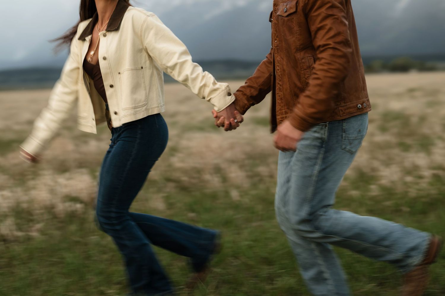 Couple holding hands running through a field, wearing jeans and jackets on a cloudy day.