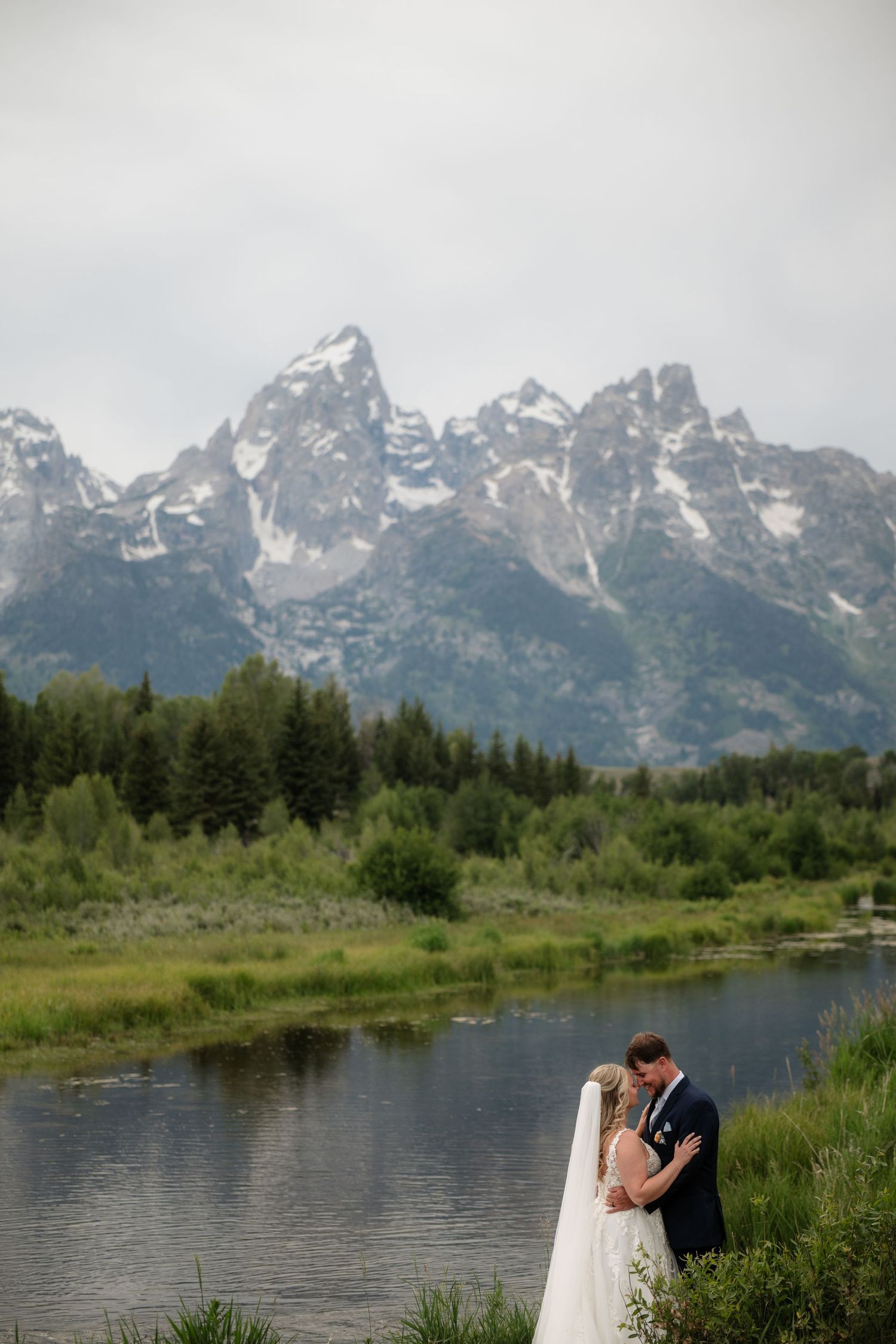 Bride and groom embrace near a serene river with the snow-capped Grand Teton mountains and lush forest backdrop.