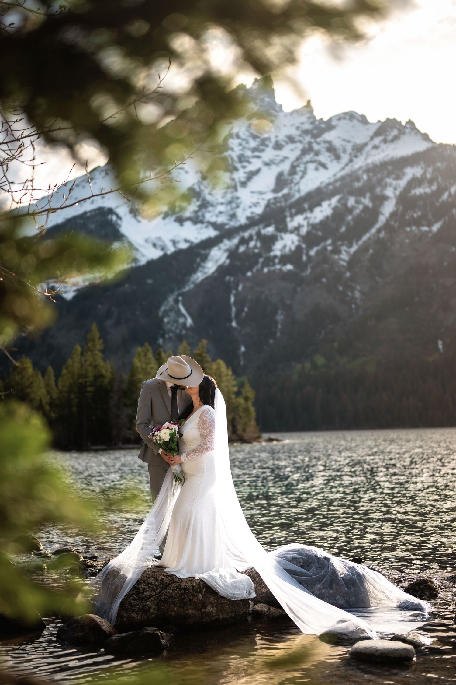 Bride and groom kissing on rocks by a mountain lake with snowy peaks and pine trees in the background.