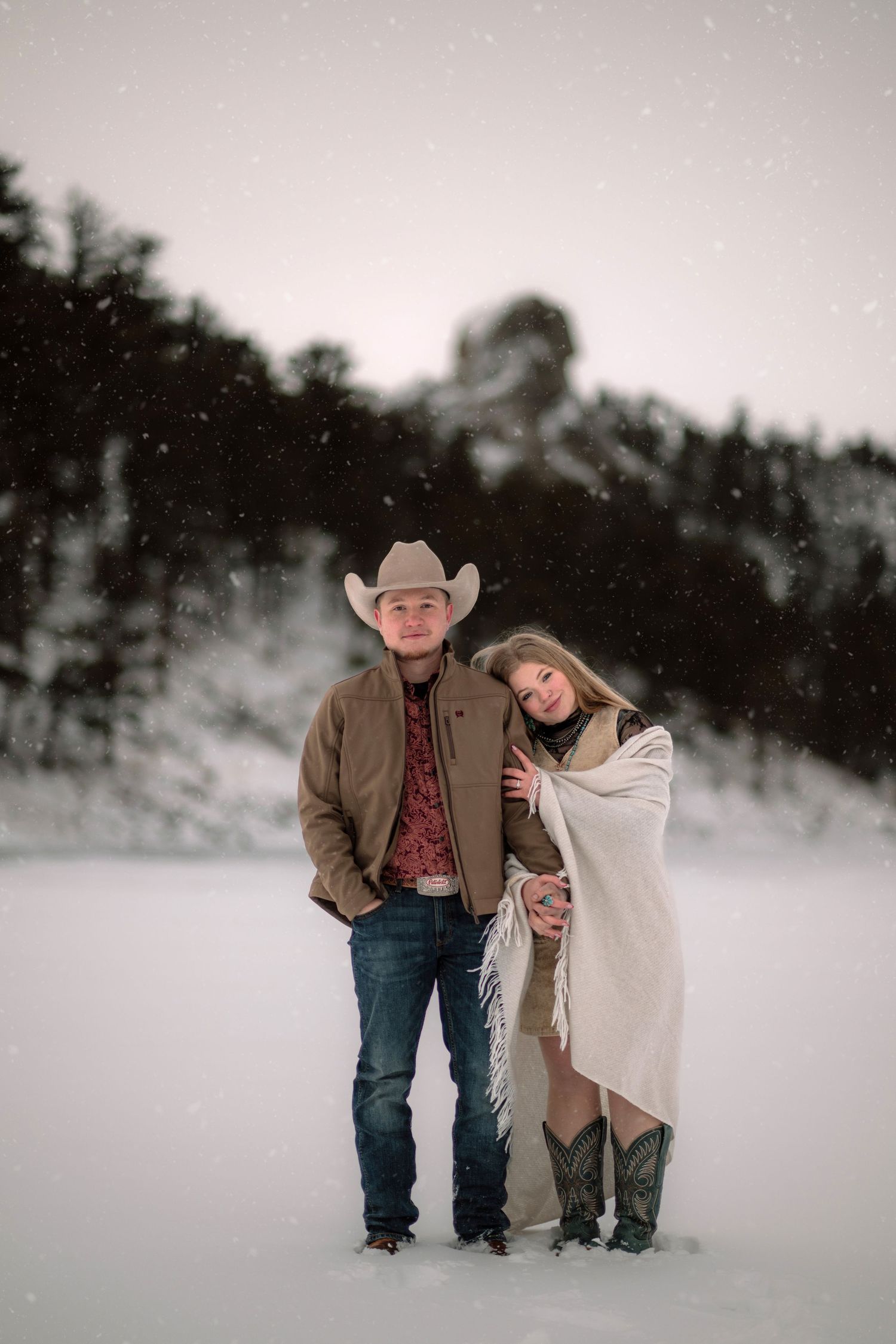 A couple standing in a snowy landscape with a mountain range in the background.