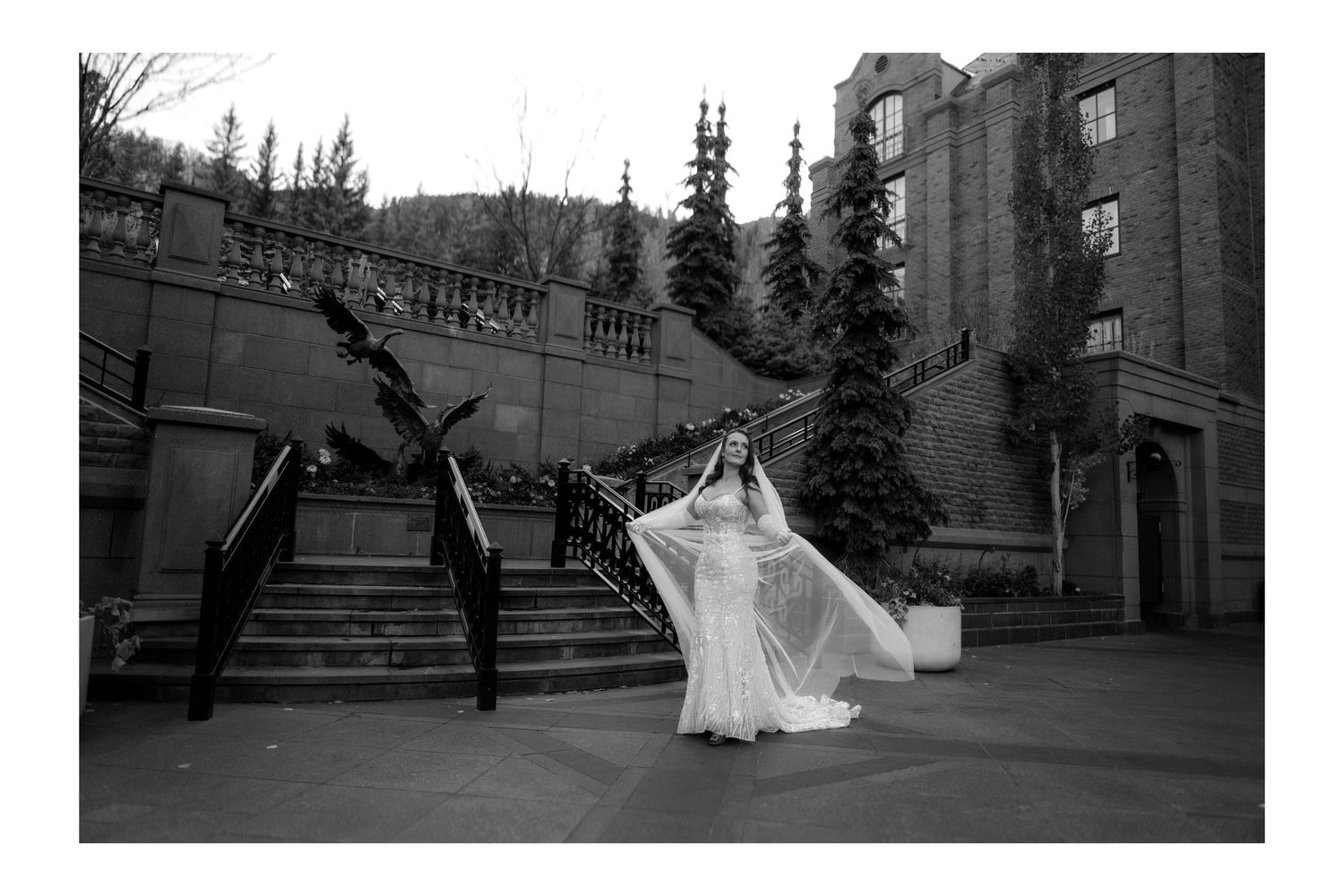 Bride in flowing white gown with veil caught in wind on grand staircase of historic stone venue in black and white.