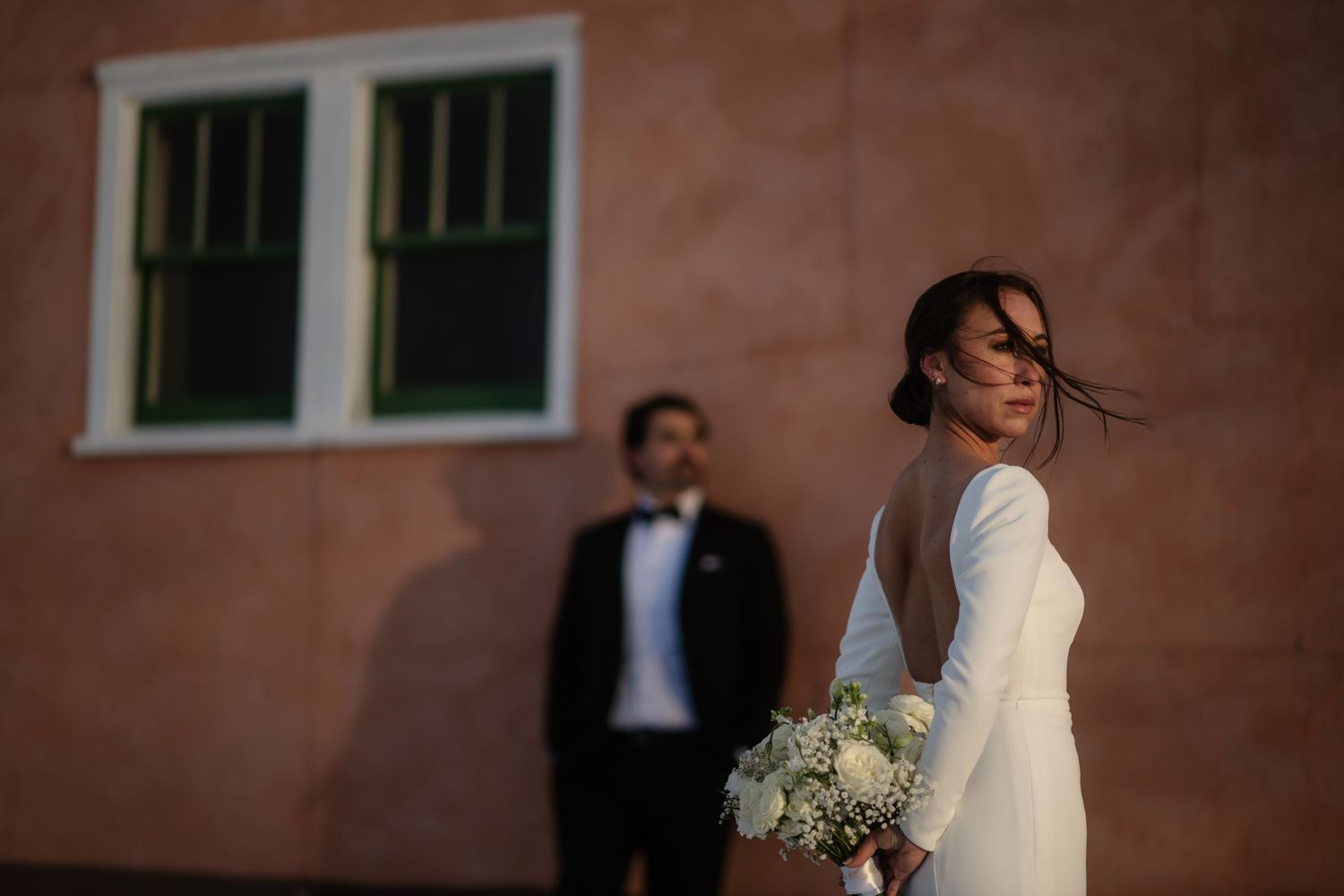 Bride in elegant backless gown holds white bouquet with groom in tuxedo against warm adobe wall.