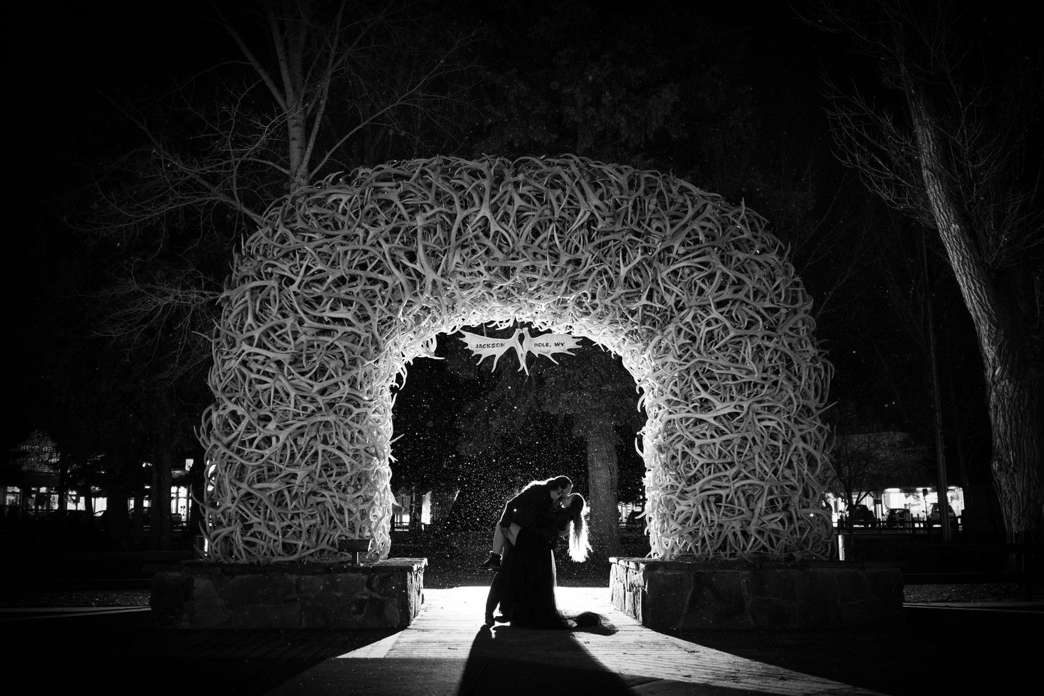 Couple silhouetted under illuminated elk antler arch at night in black and white wedding photo.