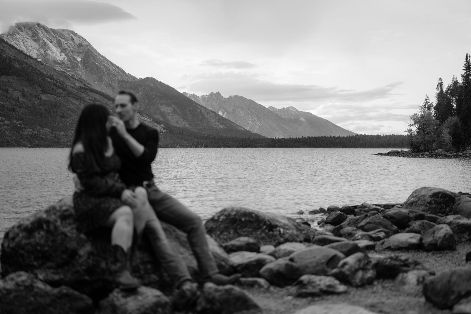 A black and white image shows a couple embracing on a rocky shore with mountains in the background.