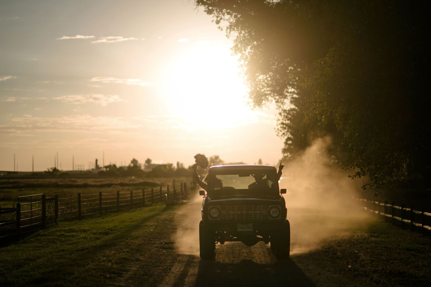 A dusty jeep drives down a rural dirt road at golden sunset, silhouetted against warm light near a tree-lined fence.