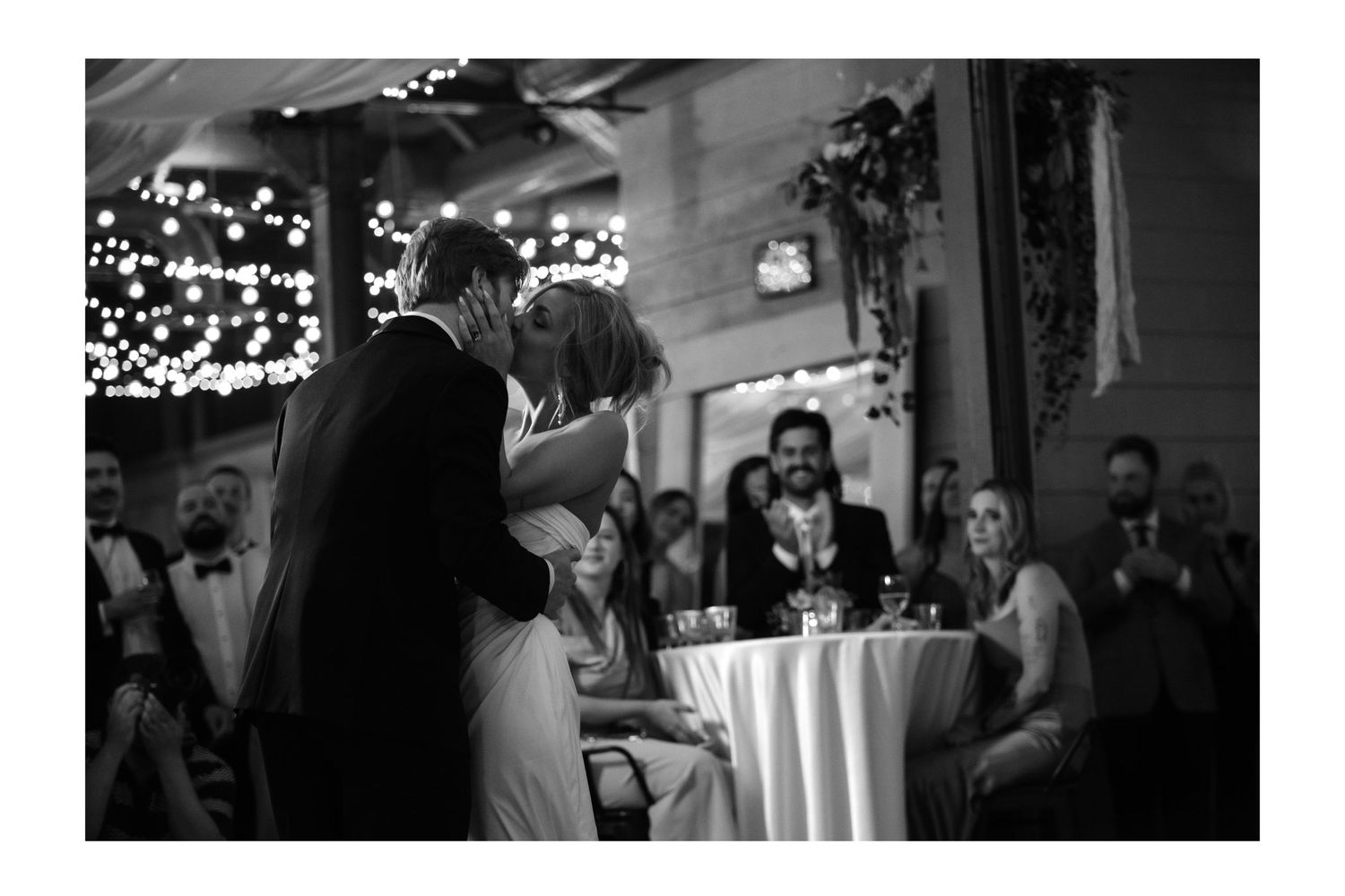 Bride and groom share first kiss at wedding reception while guests celebrate in background.