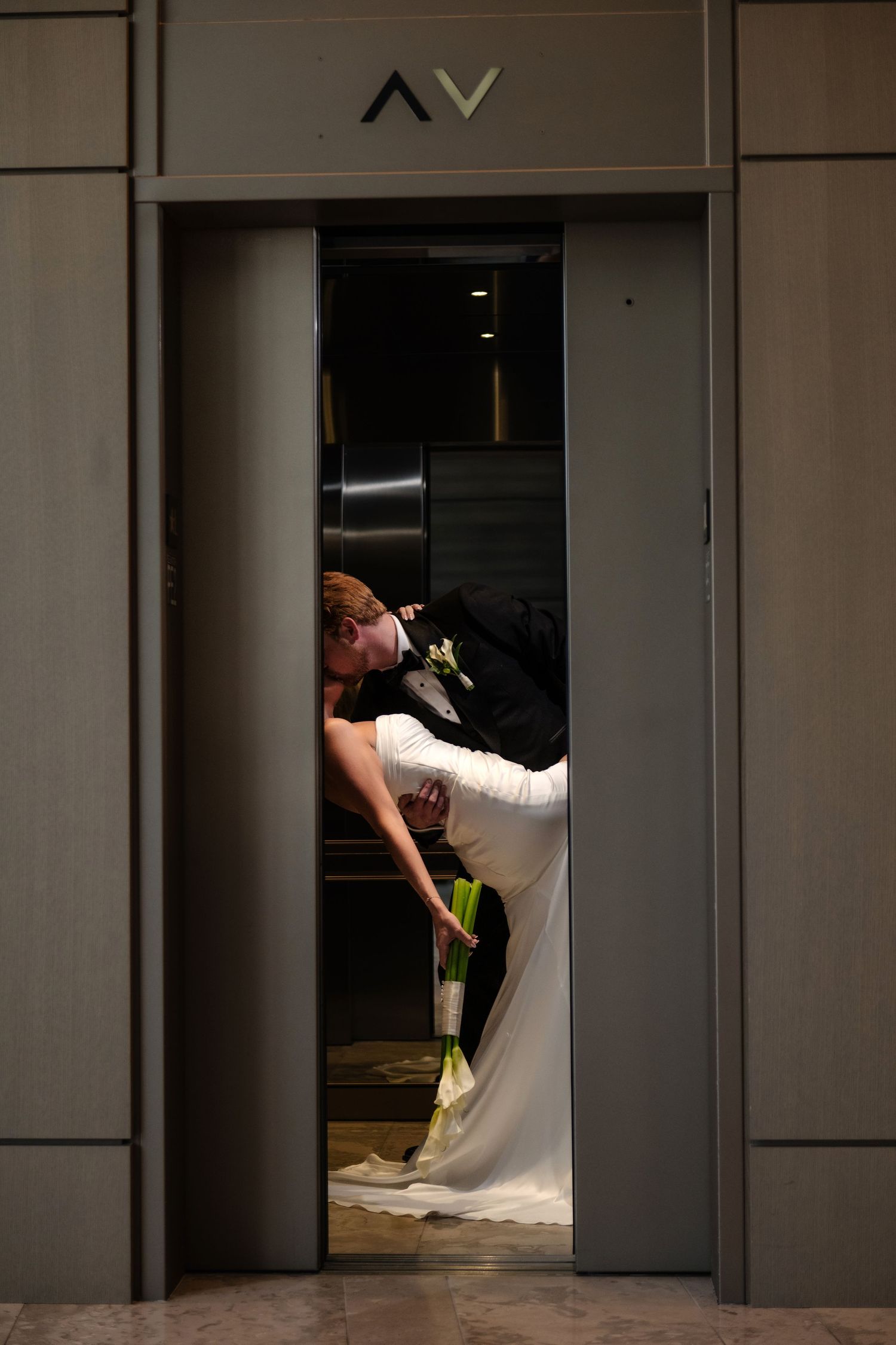 Bride and groom sharing a romantic kiss inside an elevator, bride in white gown holding yellow flowers.