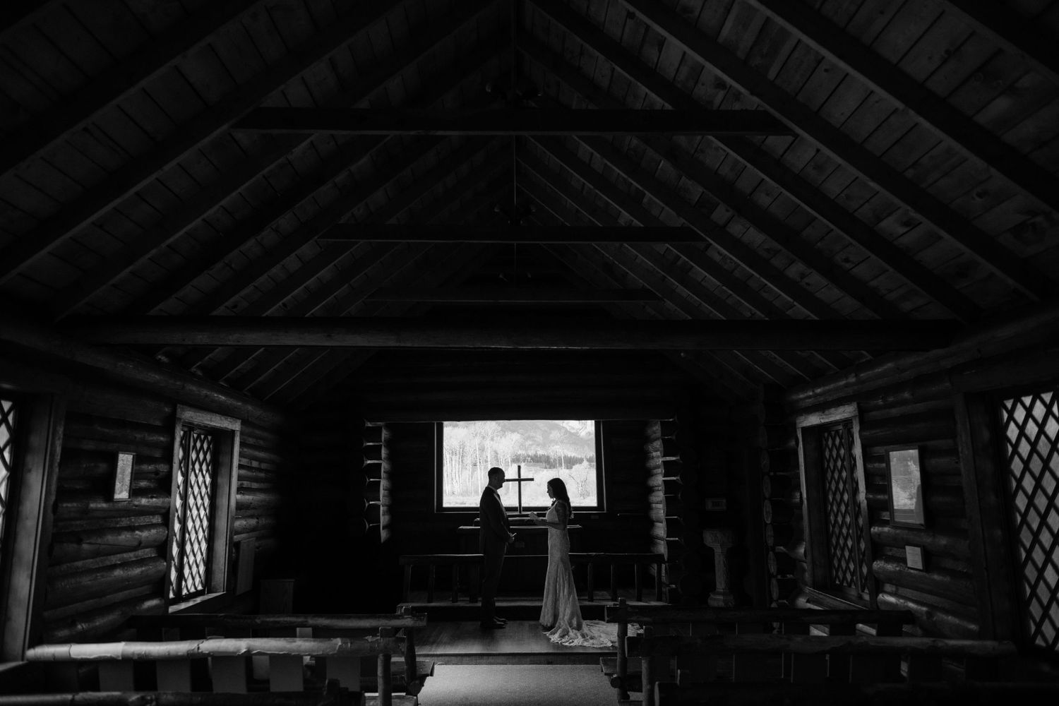 Black and white photo of a bride and groom silhouetted in a rustic log chapel with wooden pews and vaulted ceiling.