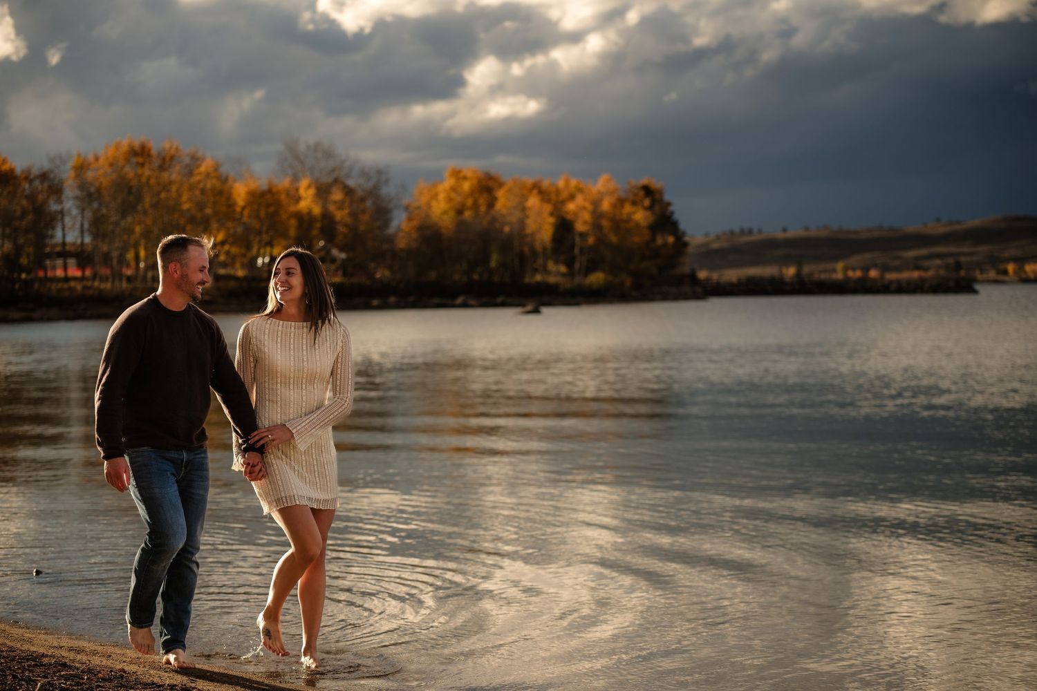 A couple strolls along the shore of a lake, with a scenic autumn landscape and a cloudy sky in the background.