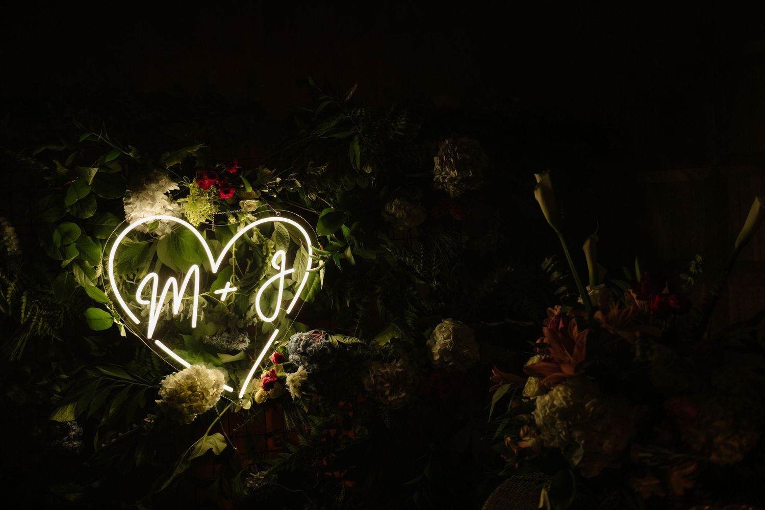 Glowing neon heart sign with initials M and J surrounded by lush floral arrangement in dark romantic setting.