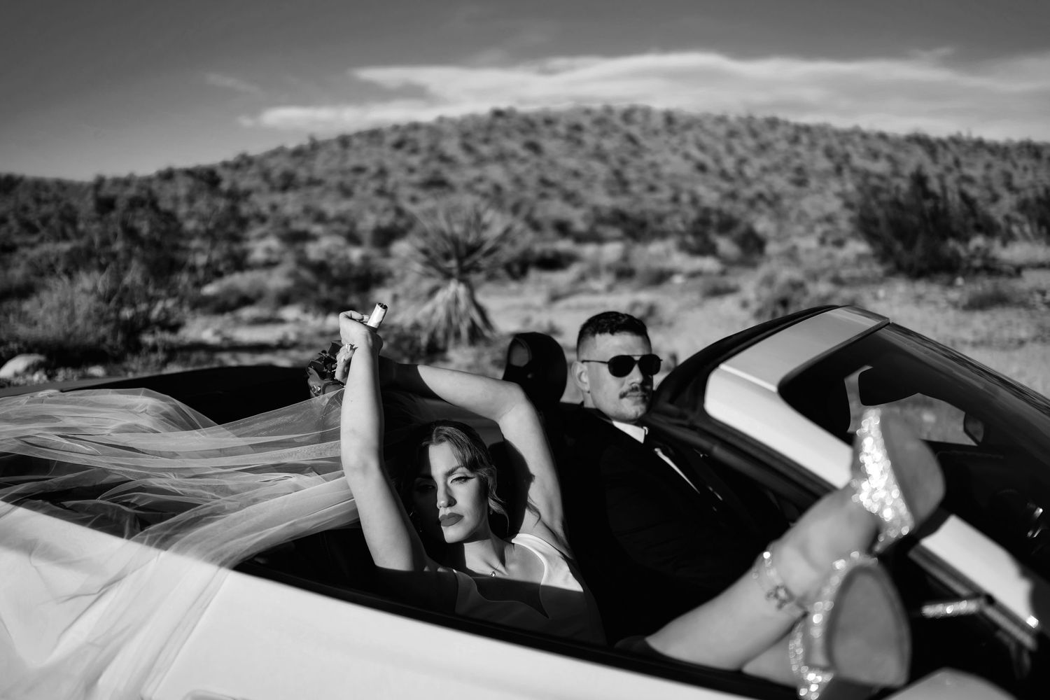 Black and white photo of a bride and groom relaxing in a convertible car in a desert landscape.