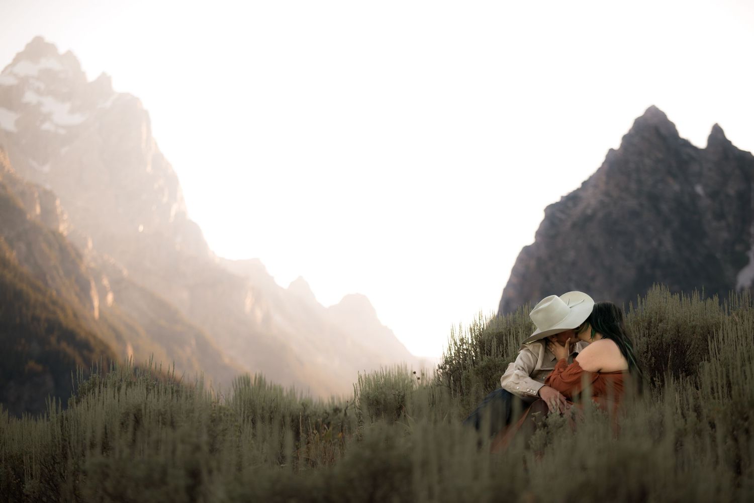 Couple sharing a romantic kiss in a lush meadow with dramatic mountain peaks in the background.