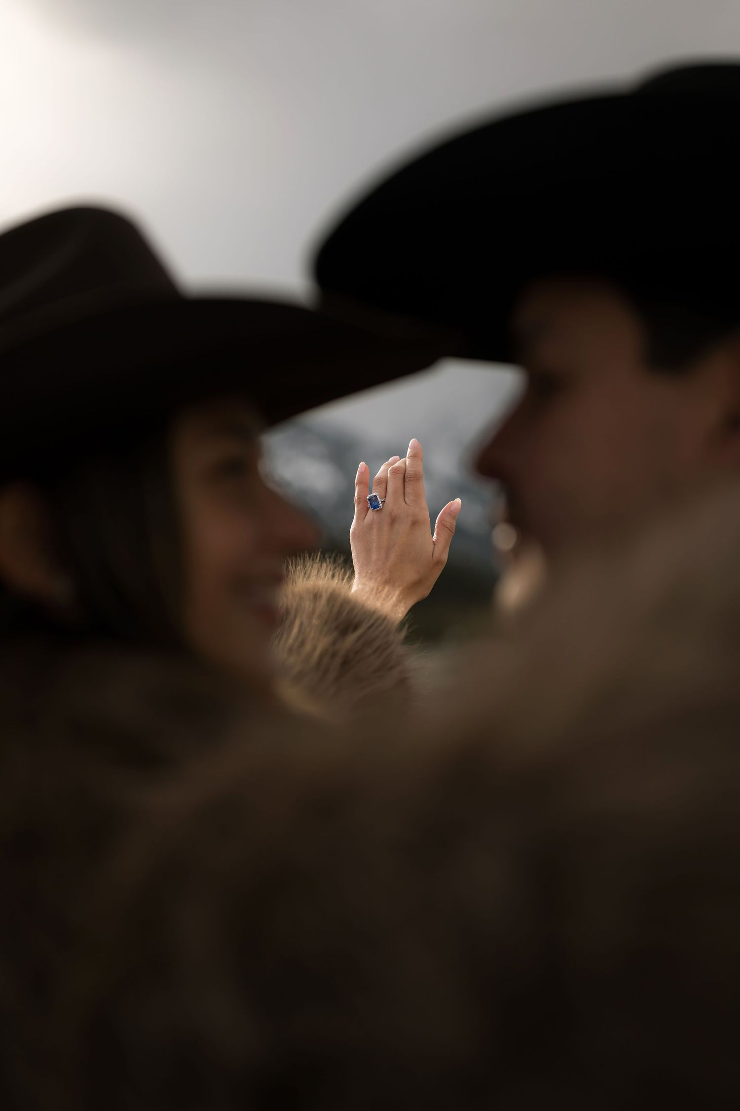 Couple in cowboy hats sharing a kiss while showing off an engagement ring with a blue gemstone stone.