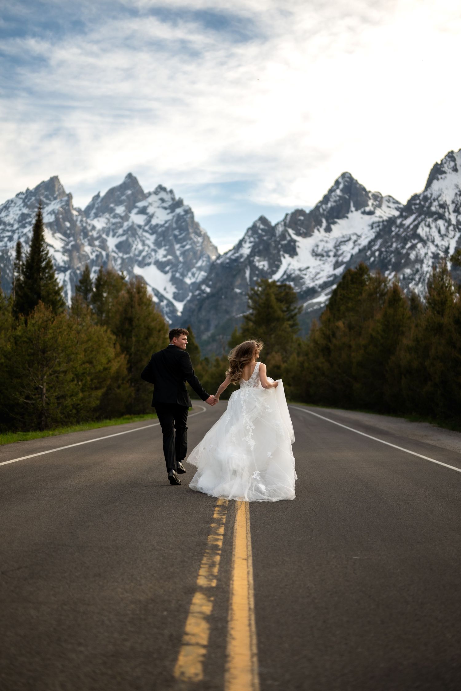 Bride in white gown and groom in black suit run hand in hand down a road with snowy Grand Teton peaks behind.