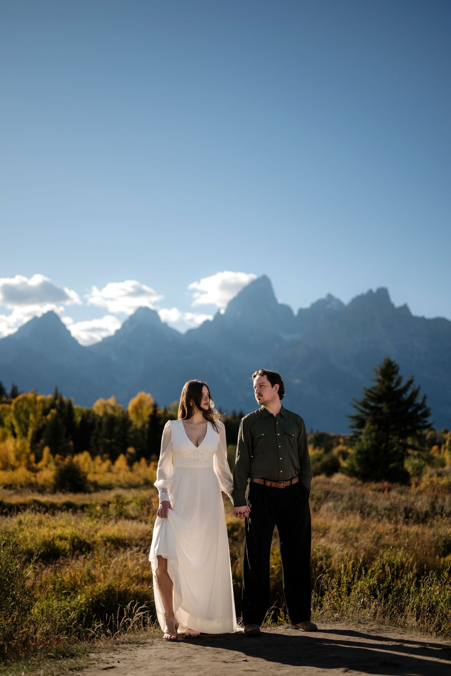 Couple holding hands in white dress and green shirt with Grand Teton mountains and fall foliage backdrop.