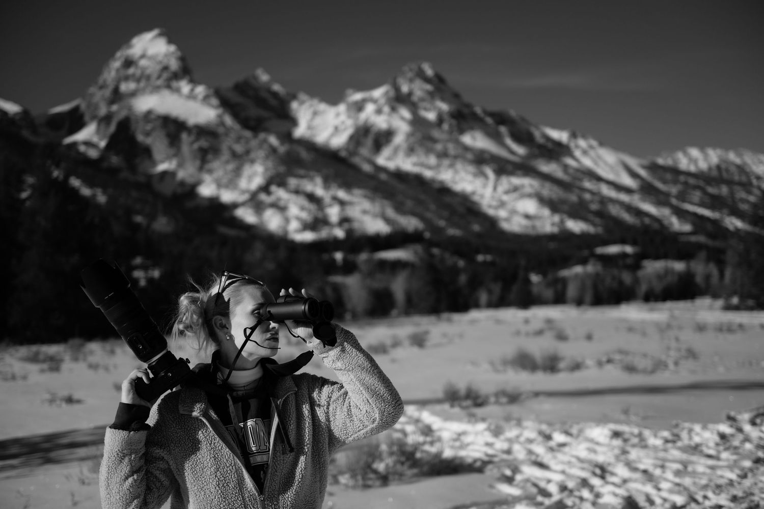 Black and white photo of a photographer with camera raised, snowy Grand Teton mountains in the background.