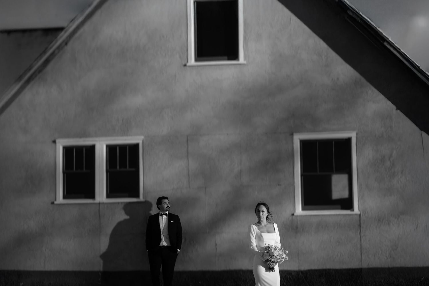 Black and white wedding photo of bride and groom standing apart against a rustic building wall with windows. Jackson Hole weddings