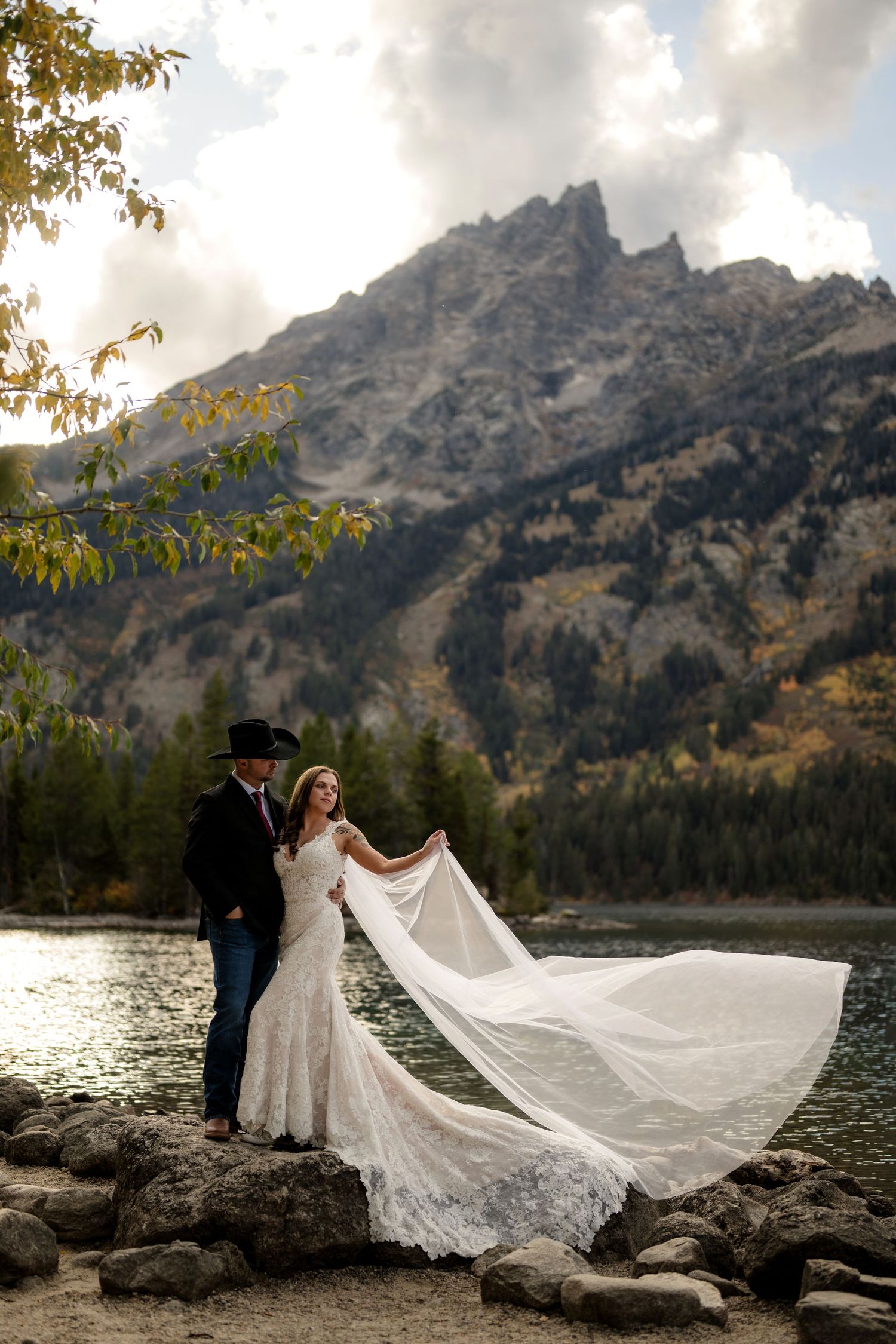 Bride in lace mermaid gown with flowing veil stands with groom by mountain lake in Grand Tetons fall scenery.