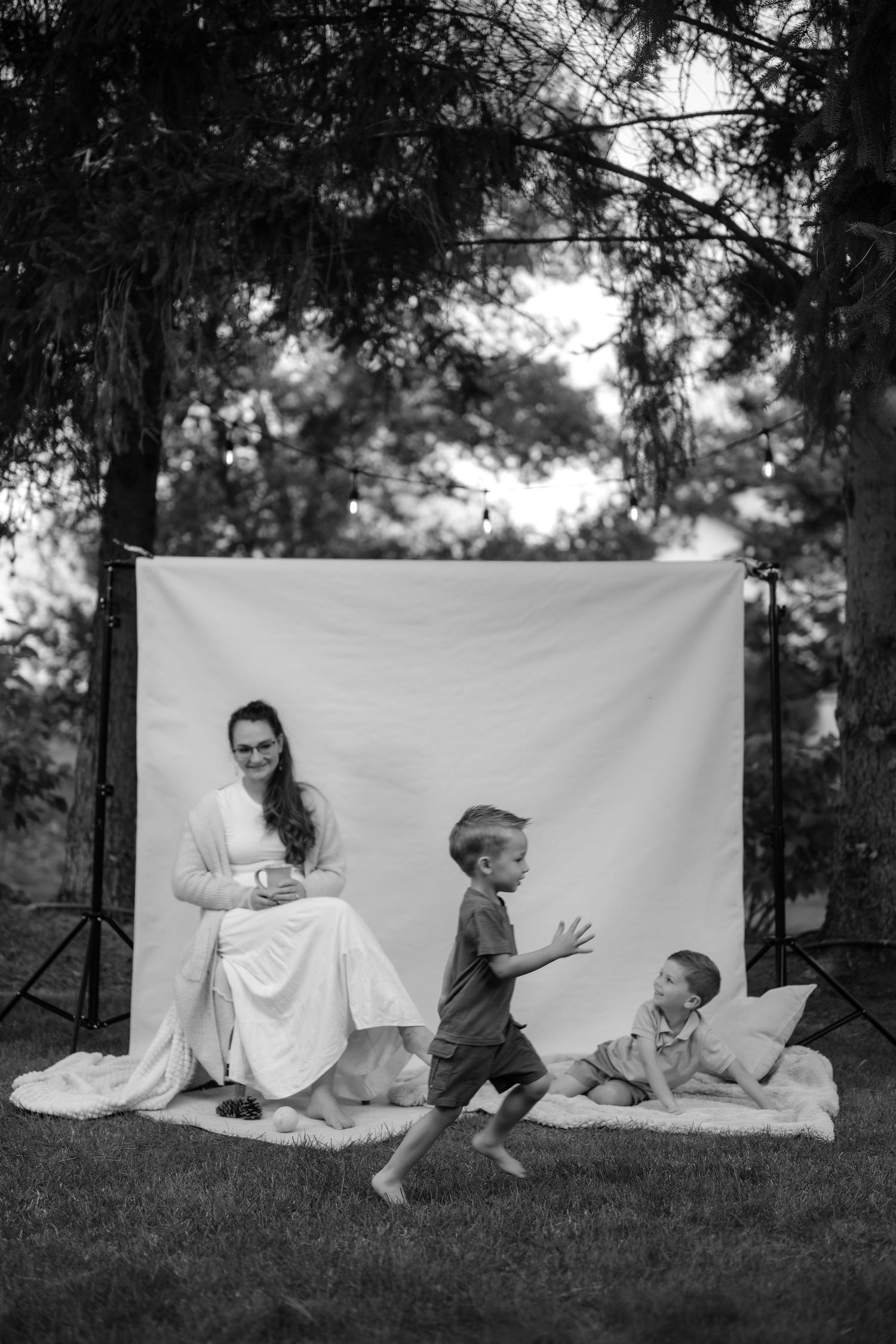 Black and white outdoor photo of a woman in white dress sitting while two young boys play on a blanket with a white backdrop.