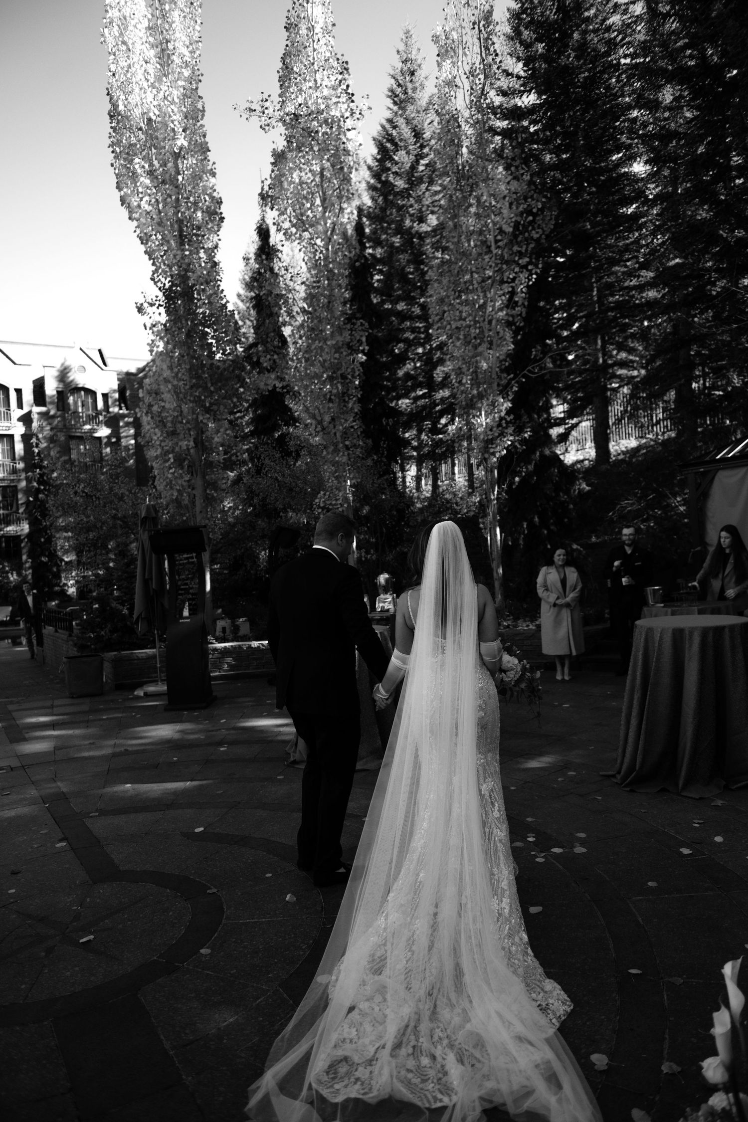 Black and white photo of bride and groom walking hand in hand toward altar surrounded by tall trees.