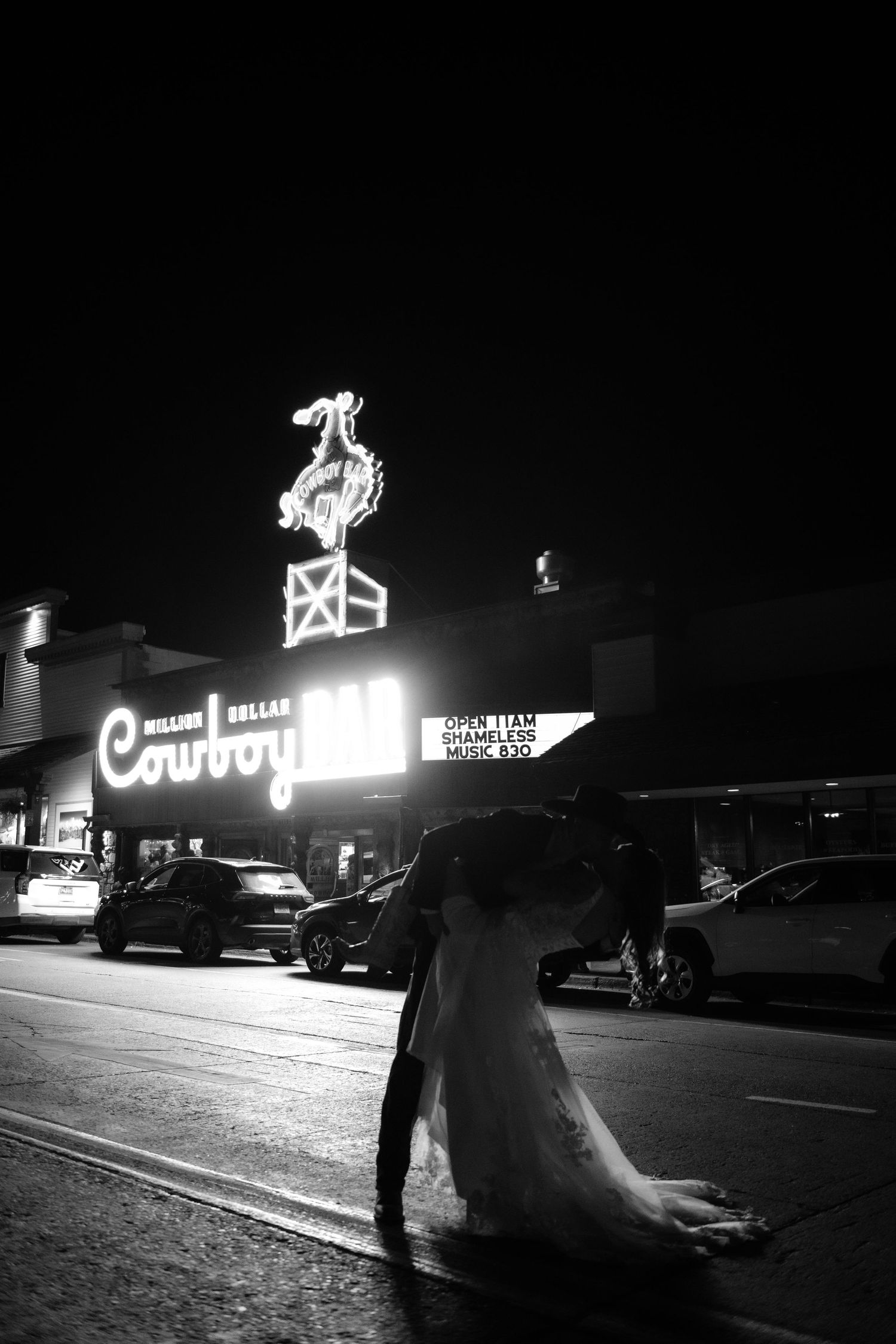 A bride in a long white dress stands on a city street at night.