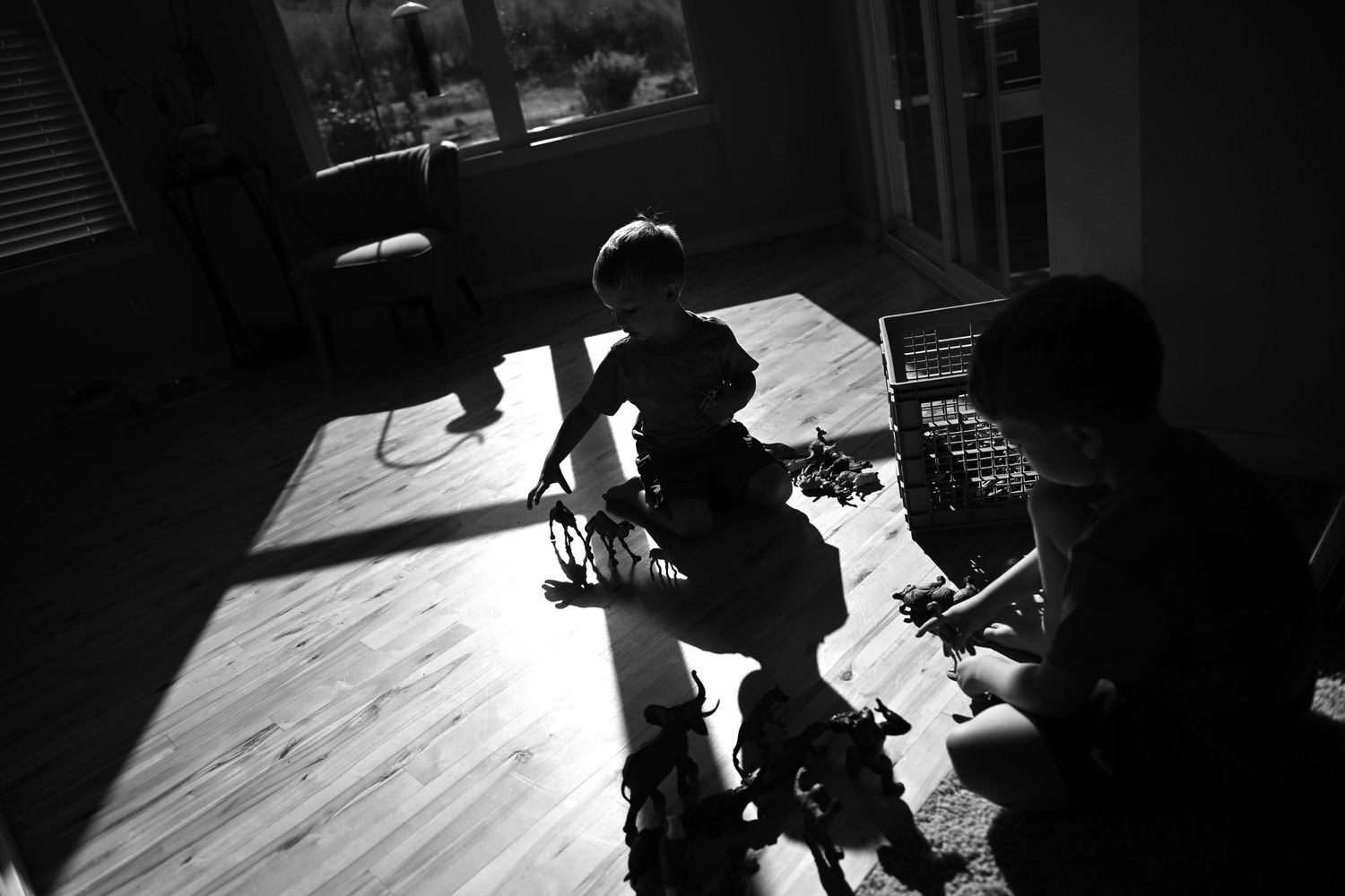 Black and white photo of two children putting on roller blades on a hardwood floor near a sunlit window.