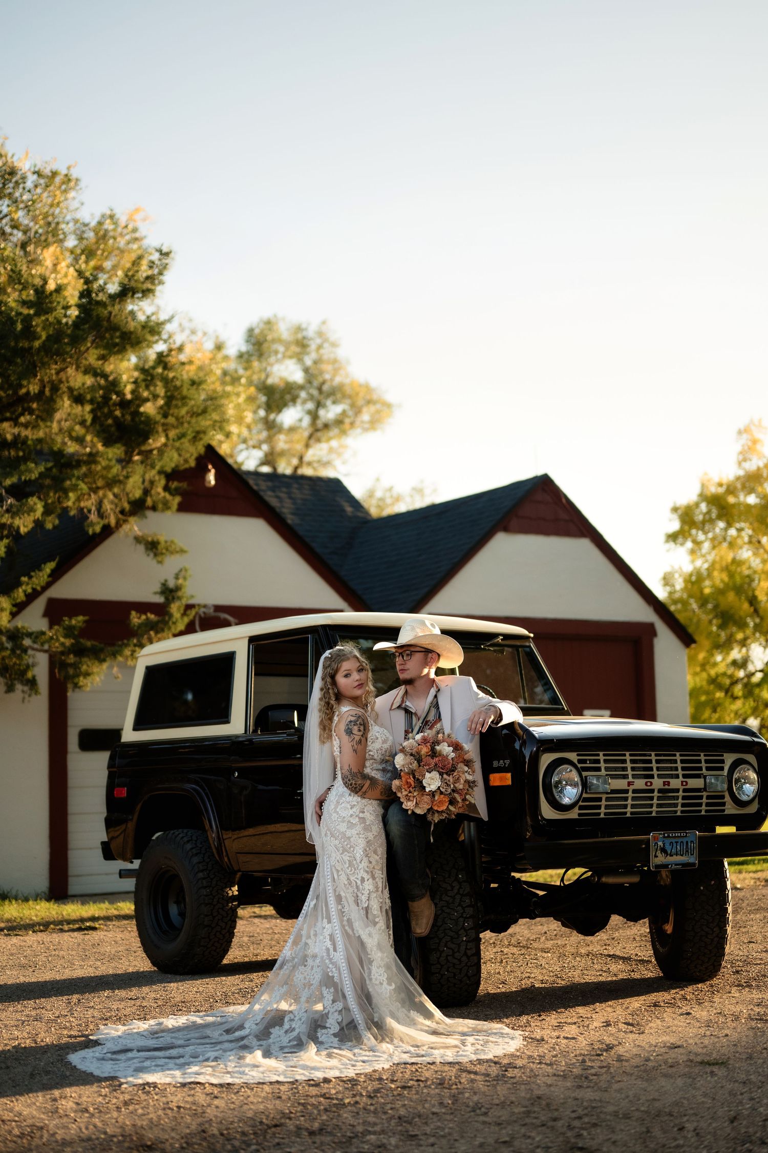 Bride and groom pose before vintage Ford Bronco at golden hour near rustic red barn on wedding day.